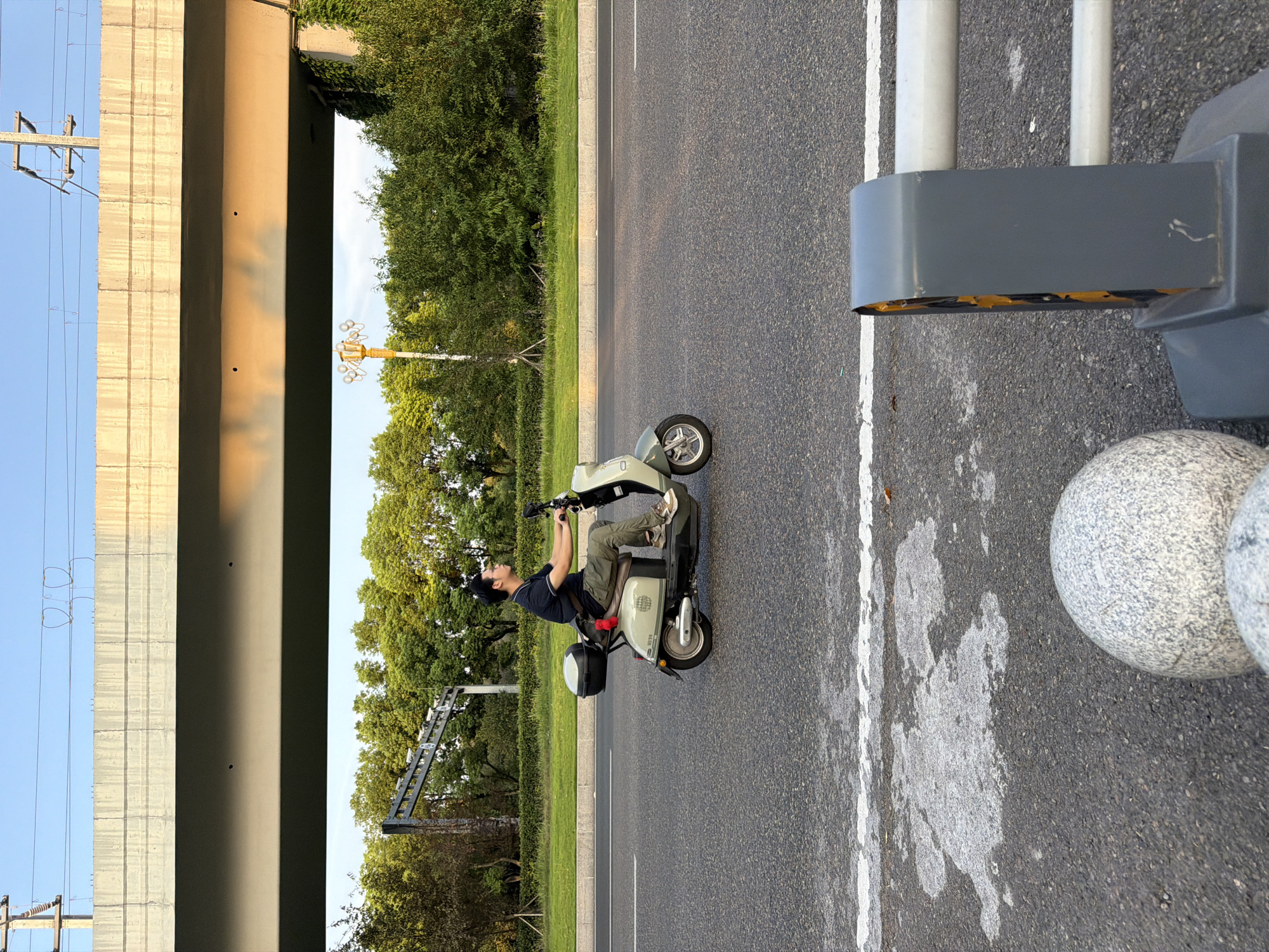 A man rides a light green electric scooter down a paved road, traveling from left to right. The man is wearing a dark blue polo shirt and olive green pants, with beige sneakers. He has short black hair and is looking straight ahead. The scooter has a black storage box on the back and a red strap hanging from the seat. Behind the scooter, a line of green trees borders the road, with a light yellow street lamp visible amongst the foliage. Above the trees, a concrete overpass casts a shadow across the scene. Overhead, power lines are strung between poles. In the foreground, the road surface is dark asphalt with white lane markings. To the right, a grey metal traffic barrier with yellow detailing stands next to two large, speckled grey boulders. The sky is clear and blue, with the warm light of sunset casting an orange glow on the underside of the overpass. The scene appears to be on a road in Jiangjin, China.