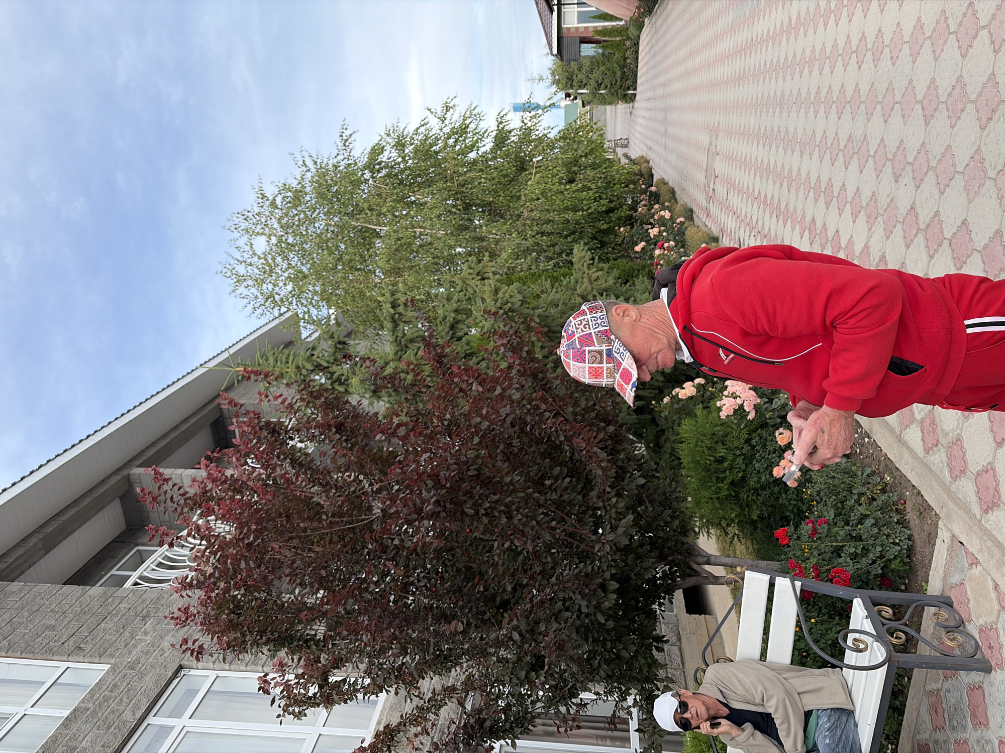 An outdoor scene depicts two men near a building and a paved walkway. On the left, a man wearing a white cap and sunglasses sits on a white bench, talking on a black cellphone. He is dressed in a light-colored jacket over a dark shirt and jeans. To his right, a man in a red hooded sweatshirt and matching pants stands, looking down at his phone. He wears a colorful baseball cap. Behind the men, a building with grey brick siding and white windows stands tall. Red and green trees flank the building, with a row of blooming pink roses visible along the edge of the walkway. The walkway itself is paved with light-colored tiles featuring a pink geometric pattern. The sky above is overcast with scattered clouds.