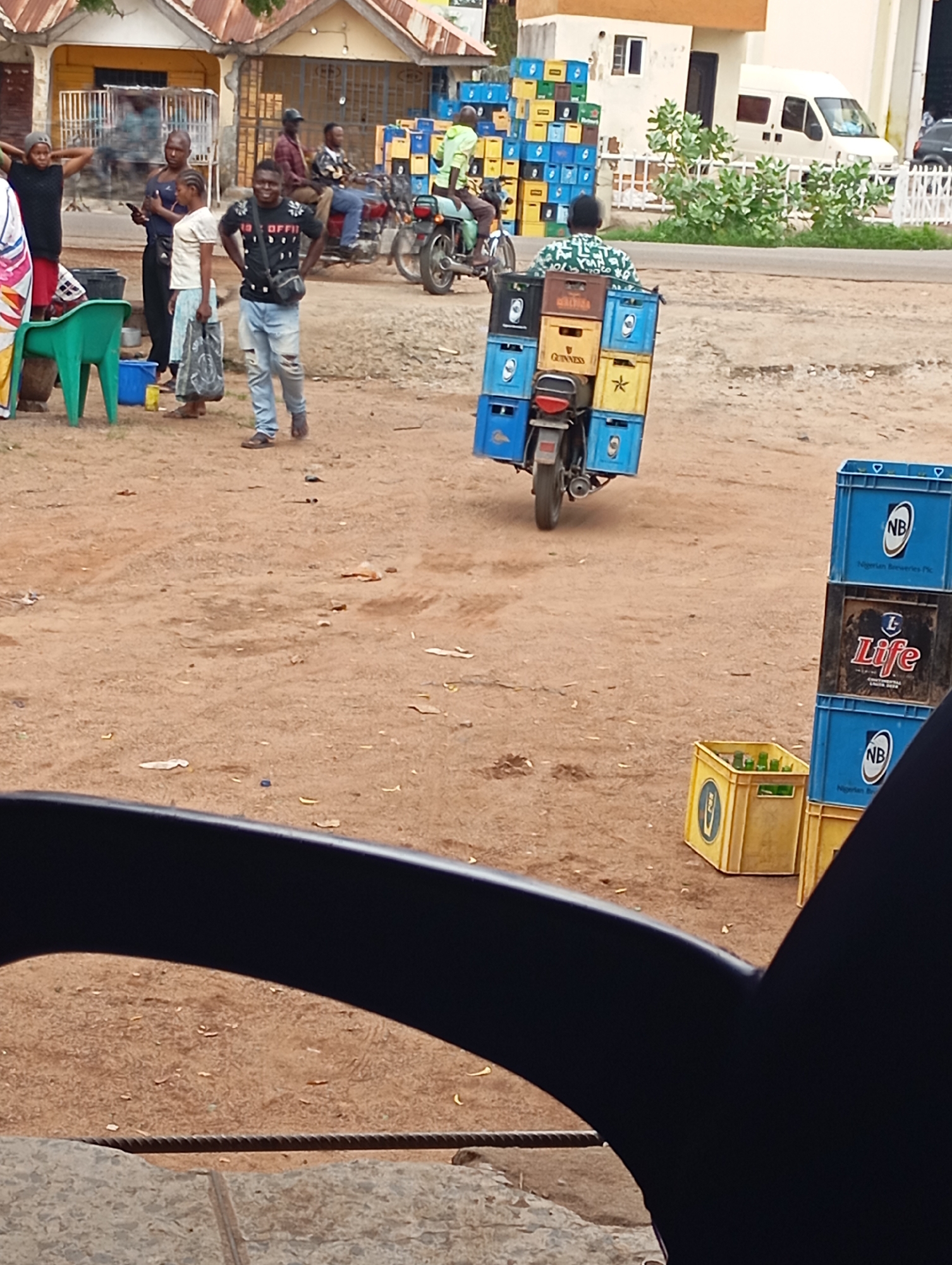 The image depicts a street scene in Shendam, Nigeria. In the foreground, a person is seated, with their body partially obscured by a dark, curved object, possibly a vehicle part. The ground is sandy and contains scattered litter. Behind this, a man on a motorcycle is carrying a load of blue and yellow crates, some labeled "Guinness." To the left, a group of people are gathered, including a man in a black t-shirt and ripped jeans, and a woman holding a white plastic bag. A green plastic chair is visible near them. In the background, buildings with corrugated iron roofs stand, and a white van is parked. Stacks of blue and yellow crates, similar to those on the motorcycle, are visible near the buildings. The overall atmosphere suggests a commercial or market area.
