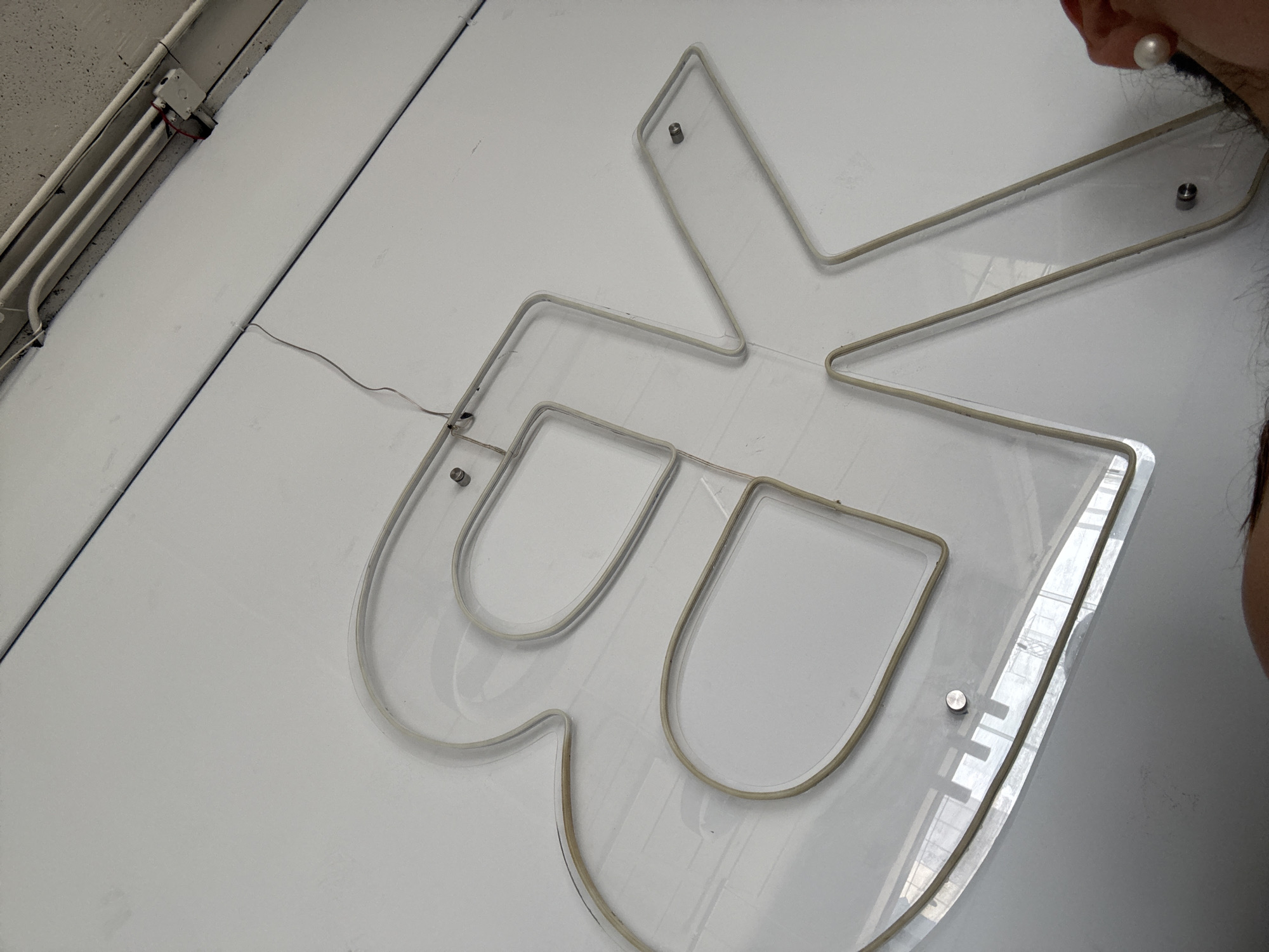 A close-up, slightly angled shot shows an illuminated "B" logo attached to a white wall. The logo appears to be constructed from clear acrylic or similar material, with the neon or LED lighting outlining the letter's shape. The "B" is affixed to the wall with metal standoffs. In the upper-left corner of the frame, electrical conduits are visible running along the wall. A small section of a person's face is visible on the right, including an ear wearing a pearl earring.
Based on the logo's design and the use of clear materials, it is likely a design element in a modern or contemporary setting.
While specific location information is unavailable from the visual content, the mention of Puebla, Mexico, suggests the possibility that this scene may be from there, depending on context and intent.