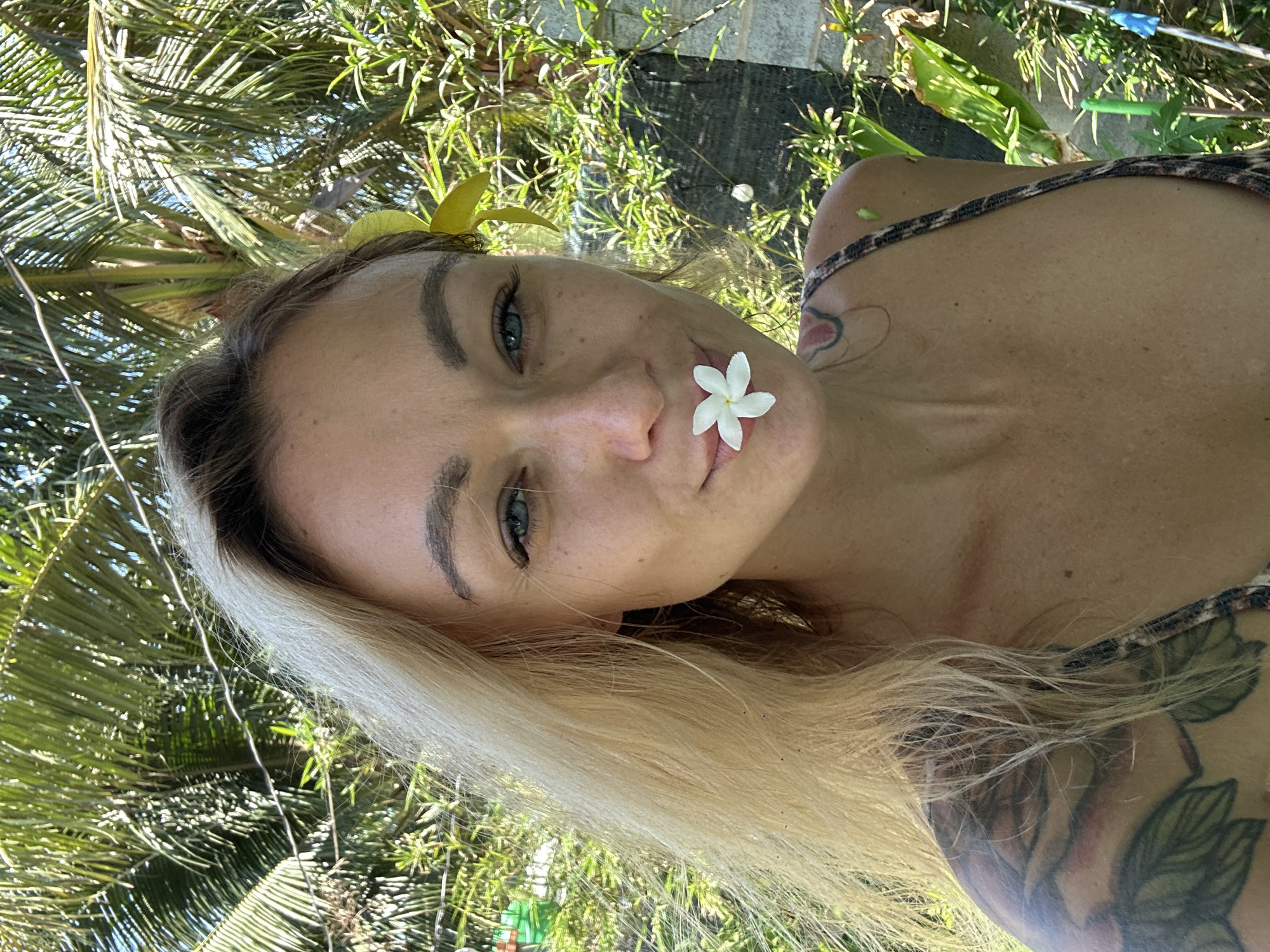 A fair-skinned woman with blonde hair is photographed outdoors with a white flower near her mouth and a yellow flower in her hair. She has fair skin, freckles, and blue eyes. Her left shoulder, as seen in the image, has a tattoo of flowers and leaves. She is wearing an animal-print style top. The background consists of foliage, including what appears to be a palm tree. The setting is suggested to be Mũi Né, Vietnam. The photo is taken at an angle, with the camera positioned to the side.