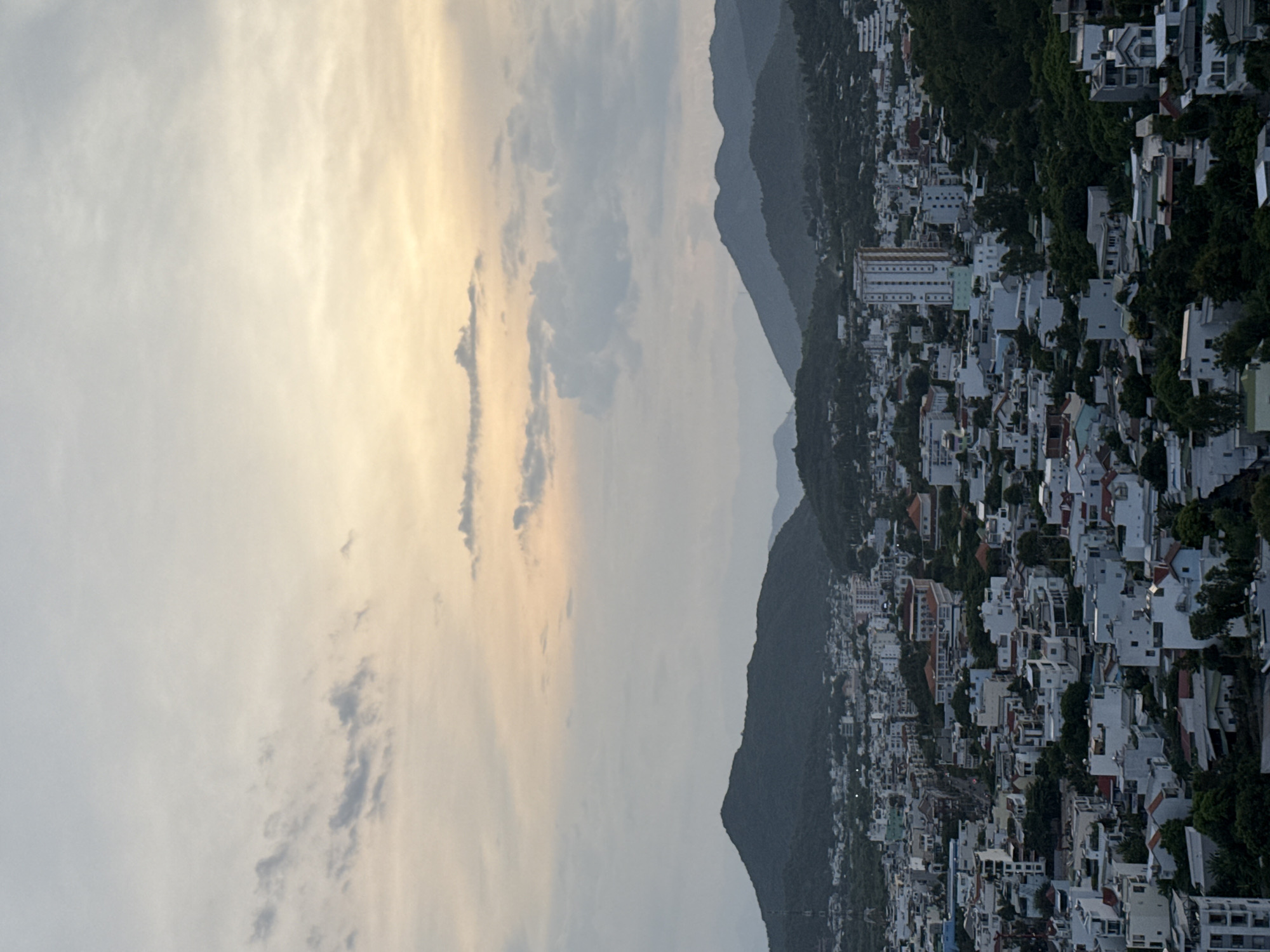 The image depicts a wide-angle aerial view of a coastal city with mountains in the background, under a hazy sky with a soft glow of sunset or sunrise. The foreground is dominated by a dense urban landscape characterized by a multitude of low-rise buildings, predominantly white with red or terracotta roofs, interspersed with lush green foliage. A prominent multi-story white building with many windows stands out in the mid-ground. The city appears to spread towards rolling, tree-covered hills and mountains that form the backdrop, with darker silhouettes against the lighter sky. The lighting suggests it is either early morning or late afternoon, casting a muted, diffused light over the scene. The location is identified as Bac Natural Heritage Area Trang, Vietnam.