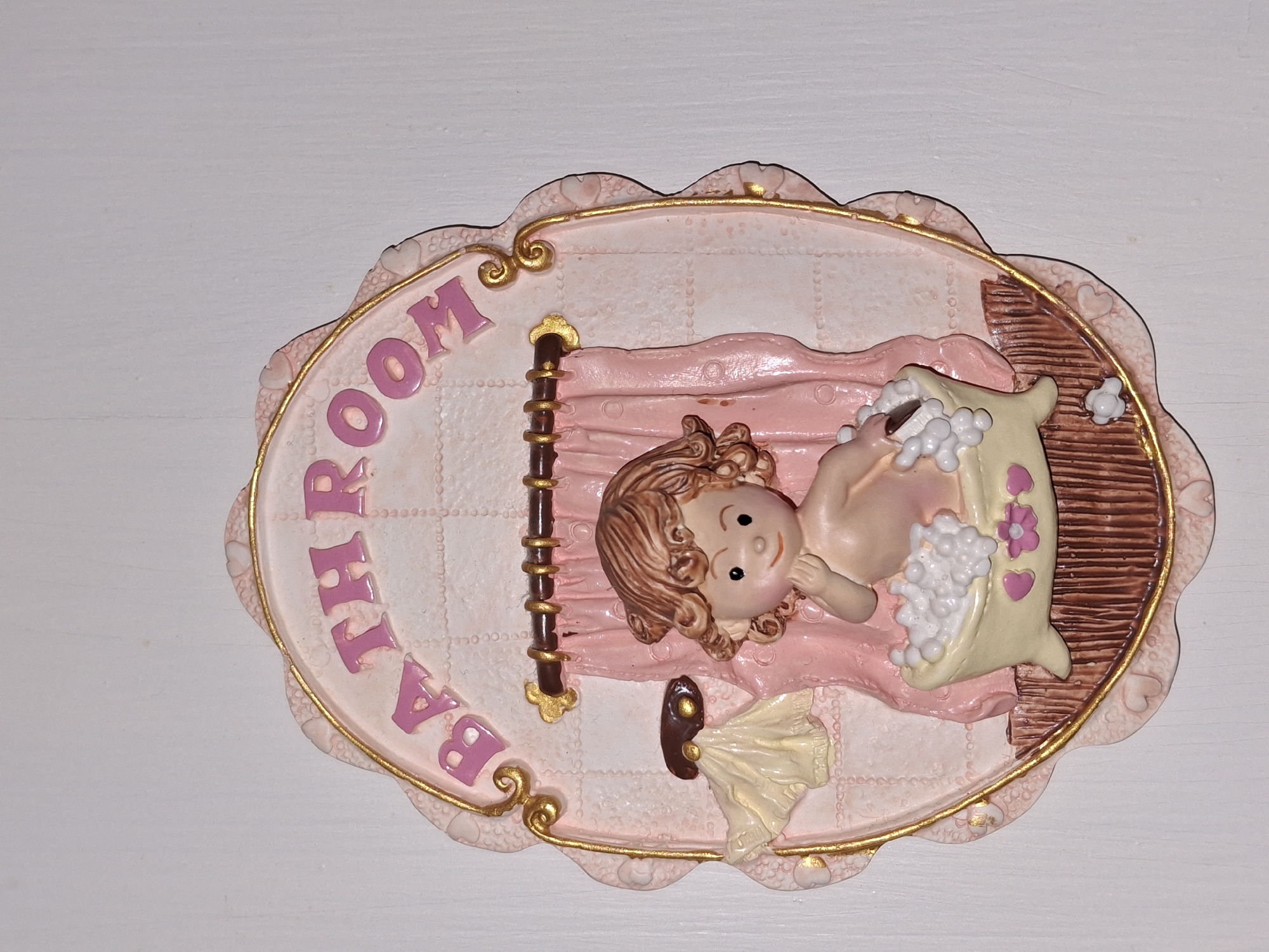 A decorative plaque reads "BATHROOM" in pink lettering against a light pink background. The plaque depicts a young child sitting in a bathtub filled with white bubbles. The child has curly brown hair and is reaching a hand towards their mouth. Pink curtains hang behind the bathtub, and a small yellow towel is draped over a hook on the left side of the plaque. The plaque is framed with a scalloped pink edge and gold trim. The background appears to be a white wall. The image is a close-up of the plaque, and no specific location in Åsgårdstrand, Norway, is visible.