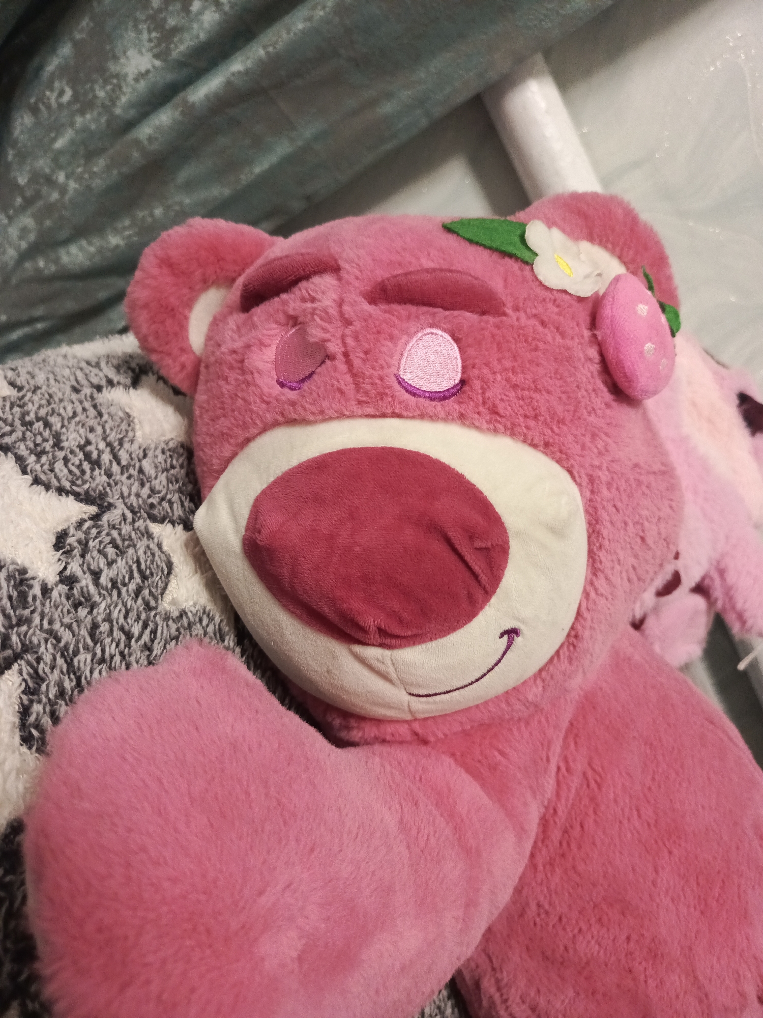 A close-up, slightly angled shot shows a plush toy bear, Lotso Huggin' Bear from Toy Story, lying down. The bear is a bright pink color and appears to be made of soft, fuzzy material. It has embroidered features including closed eyes with purple stitching, a light pink nose, and a smiling mouth with purple stitching. A small, white felt flower with a yellow center and a green leaf is attached to the top of its head, along with a pink strawberry-shaped accessory. The bear is resting on a gray and white patterned blanket that features star shapes. In the background, a draped teal-colored fabric can be seen. The image is well-lit, with some reflections visible on the background fabric. There is no indication of the location being in Вылково, Russia.