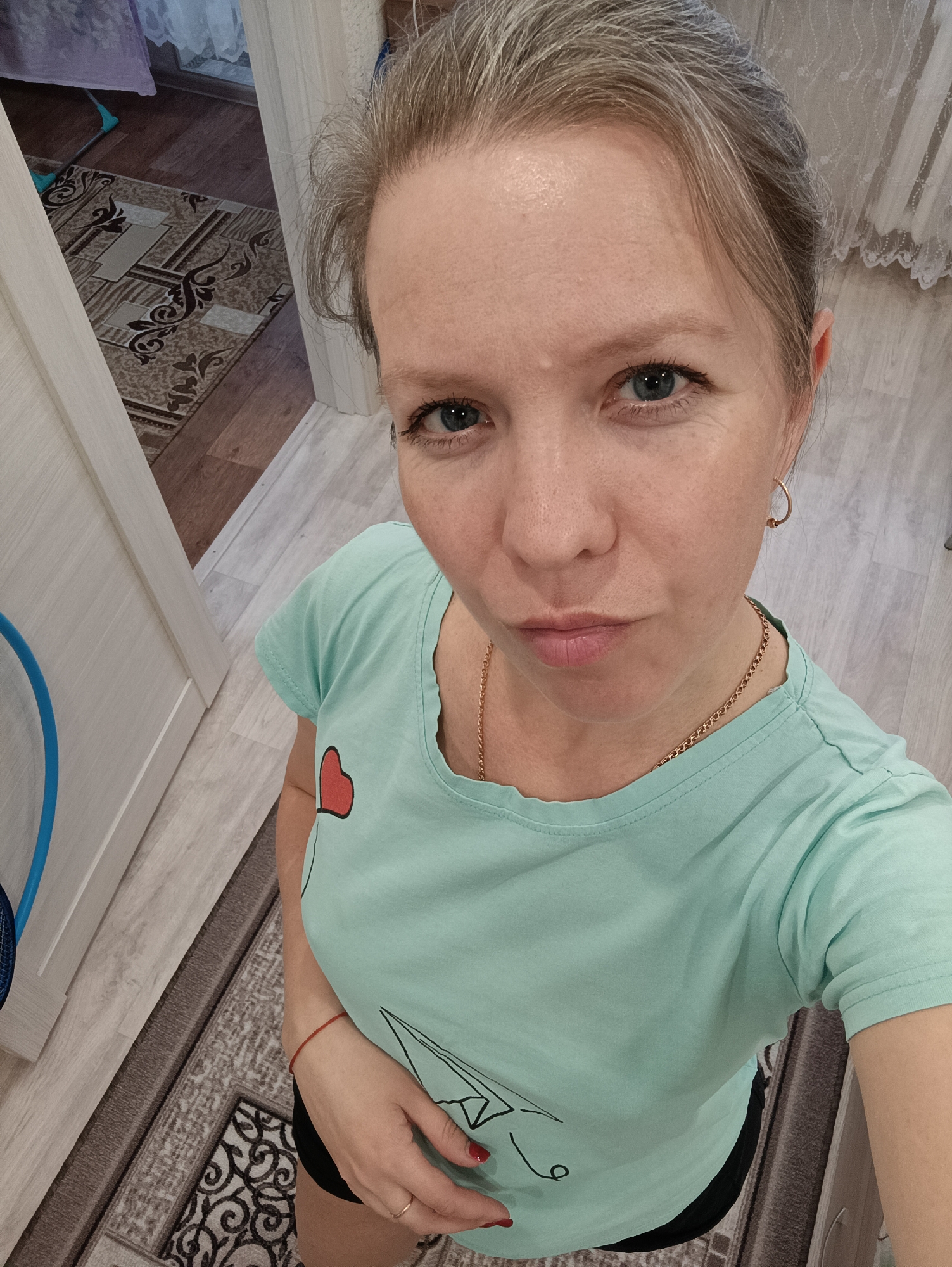 A woman takes a selfie indoors in Chaikovsky, Russia, captured from a high-angle perspective. She appears to be in her 30s or 40s, looking directly at the camera with a slight pout or serious expression. Her hair, light brown with hints of gray, is pulled back, and she has striking blue eyes. Her complexion is fair, showing natural skin texture.
She is dressed in a light mint green short-sleeved t-shirt featuring a red heart graphic on the left chest and a black line drawing, possibly a paper airplane or boat, below it. She wears black shorts or bottoms. Her accessories include small gold hoop earrings, a thin gold chain necklace, a red string bracelet on her right wrist, and a thin gold ring on her right ring finger. Her fingernails are painted red. Her right hand rests gently on her lower torso.
The background suggests a domestic setting, likely a hallway or room. The flooring comprises light-colored wood laminate with a patterned rug in shades of brown, beige, and black. A light-colored door frame is visible on the left, and a sheer, lace-patterned curtain hangs in the background on the right. Further into the background, a purple curtain or fabric and a cleaning tool can be discerned. Part of a blue object, possibly a fan or a child's toy, is visible on the far left.