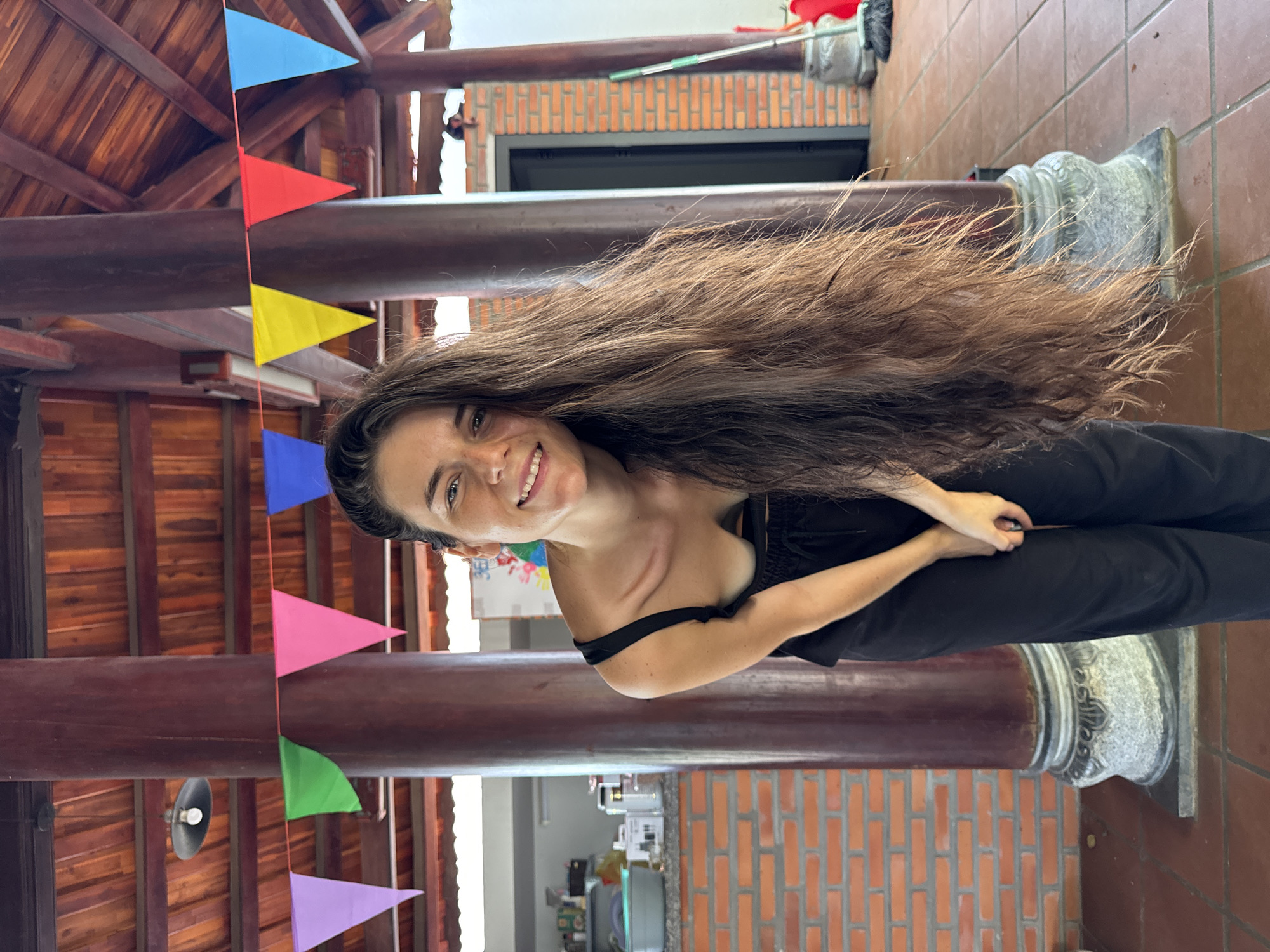 A light-skinned female individual with long, dark brown hair, wearing a black tank top and black pants, is depicted smiling directly at the viewer. She is positioned centrally, bending forward at the waist with her hands clasped in front of her. Her hair cascades over her right shoulder and chest.
The scene is an indoor or semi-open structure in Mũi Né, Vietnam, characterized by traditional Vietnamese architecture. The background features several large, dark reddish-brown cylindrical pillars with ornate gray stone bases. A string of multi-colored triangular flags (purple, green, pink, yellow, red, light blue) is strung between two pillars above the subject. The ceiling consists of dark wooden beams and rafters, with a red-tiled roof visible above. A single light fixture with a bare bulb hangs from a beam on the left.
To the left of the subject, a brick wall forms part of a utility or kitchen area, displaying a countertop with a light blue plastic tub and various white containers. To the right of the subject, another brick wall features a dark doorway, and a red bucket and a green-handled mop/broom are leaning against it. A small, dark animal figure is visible perched on a wooden beam in the distant right background. The floor is covered with reddish-brown tiles. The lighting appears to be natural, casting soft shadows.