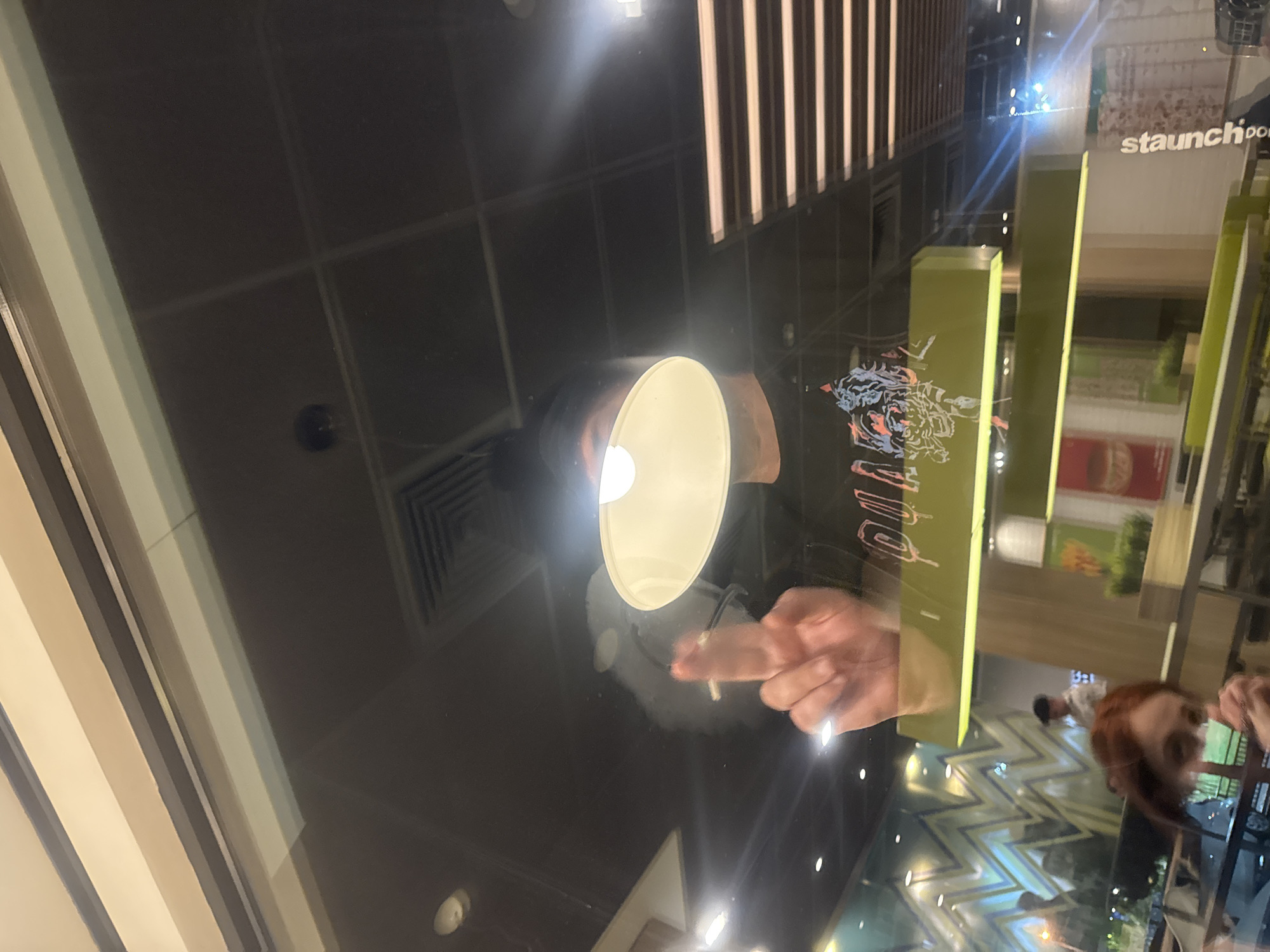 The image appears to be a reflection on a glass table in a commercial establishment, possibly a restaurant or cafe. A lit lamp hangs above the table. Visible in the reflection is a hand holding a lit cigarette, with smoke emanating from it. The reflection also shows part of the restaurant's interior, including signage with the name "staunch." The location is likely in Tbilisi, Georgia. A person with red hair is visible in the background reflection. The floor has a zig-zag pattern.