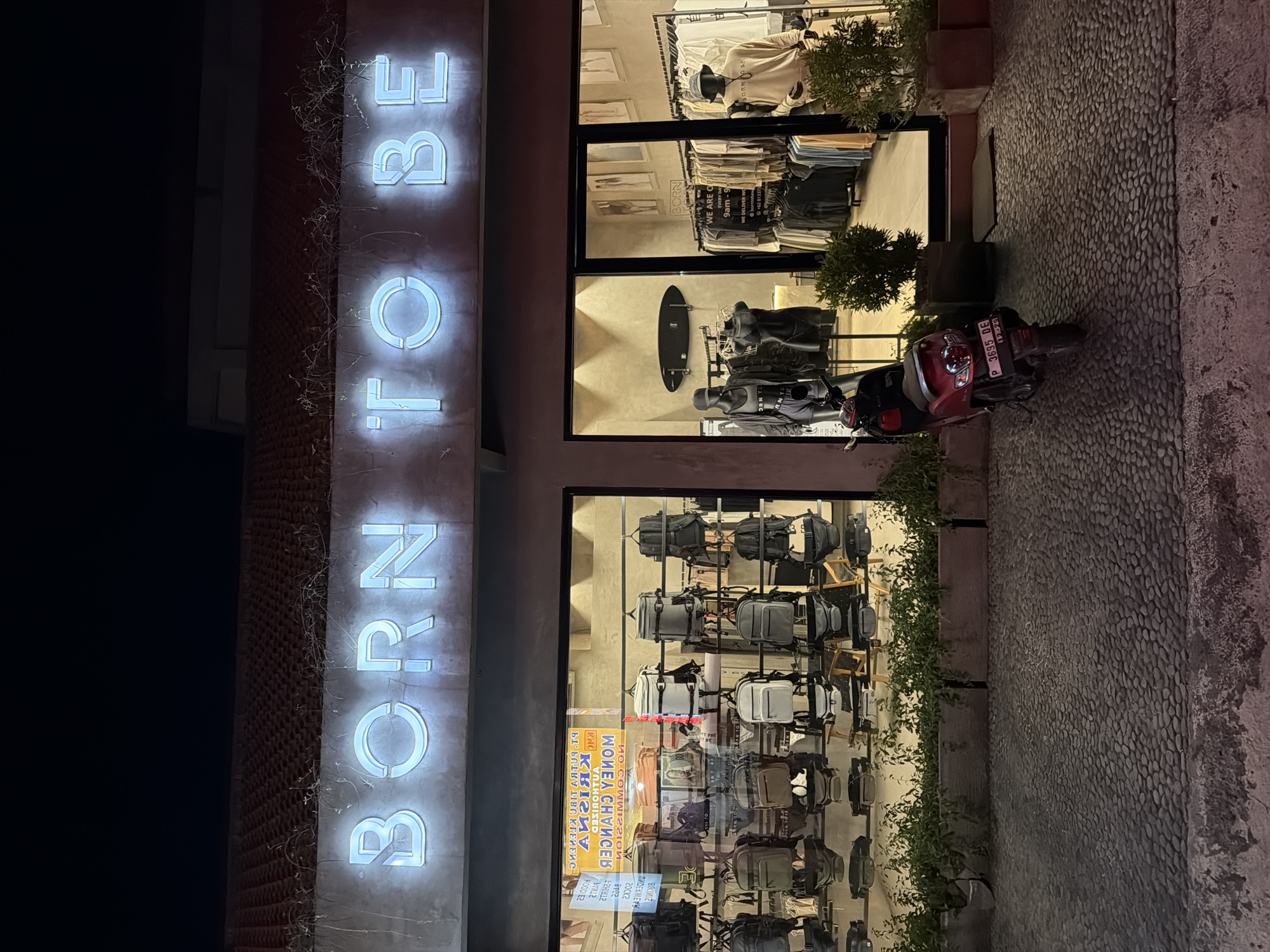 The image shows the facade of a store called "BORN TO BE" in North Kuta, Indonesia. The store has a vertical sign with the name in illuminated letters. The store has several windows displaying its merchandise, which includes clothes, surfboards, and backpacks. A red scooter is parked on the cobblestone sidewalk in front of the store. Plants are visible along the edge of the building.