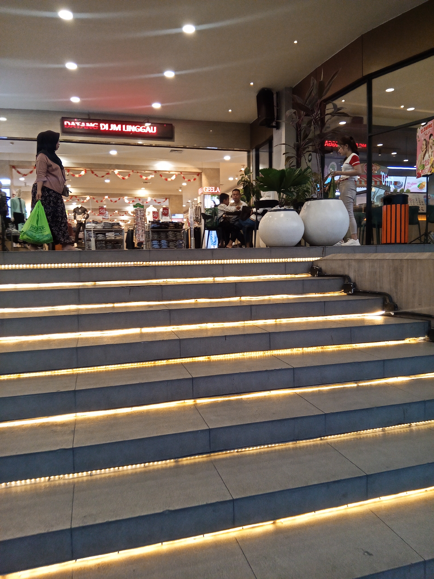 The image depicts an indoor shopping environment, likely a department store, based on the displays of clothing and signage. In the foreground, a wide set of concrete stairs with integrated yellow LED lighting leads upwards. The lighting creates a warm glow on the steps.
On the landing at the top of the stairs, several individuals are visible. To the left, a woman wearing a hijab and a patterned skirt walks towards the stairs, carrying a bright green shopping bag. Behind her, a clothing display featuring mannequins and racks of garments is visible, with red and white festive decorations overhead. A red LED sign displays "DATANG DI JM LINGGAU," indicating that the location is JM Linggau in the city of Lubuklinggau, Indonesia.
Further to the right on the landing, a group of people are seated, appearing to be resting or conversing. Among them is a woman wearing a red and white striped shirt, standing and looking towards the right. Two large white decorative pots, one significantly larger than the other, are placed near this group. Beyond them, a window offers a glimpse of an exterior scene or another section of the store, with plants and more decorative elements.
The overall impression is of a well-lit and maintained commercial space, with a mix of shoppers and decorative features. The presence of the sign clearly identifies the location as JM Linggau in Lubuklinggau, Indonesia.