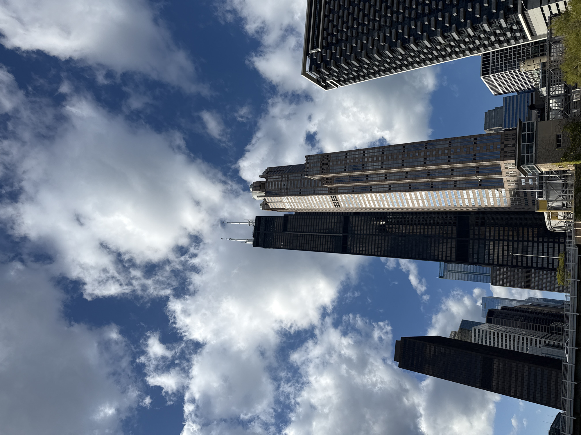 This image shows a low-angle view of several skyscrapers in Chicago, United States, against a backdrop of blue sky with scattered white clouds. The prominent buildings are the Willis Tower (formerly Sears Tower), and the Aon Center. Other unidentified buildings are visible in the background. The buildings are various shades of grey and black, with some reflecting sunlight. The perspective suggests the photo was taken from ground level, looking upwards.