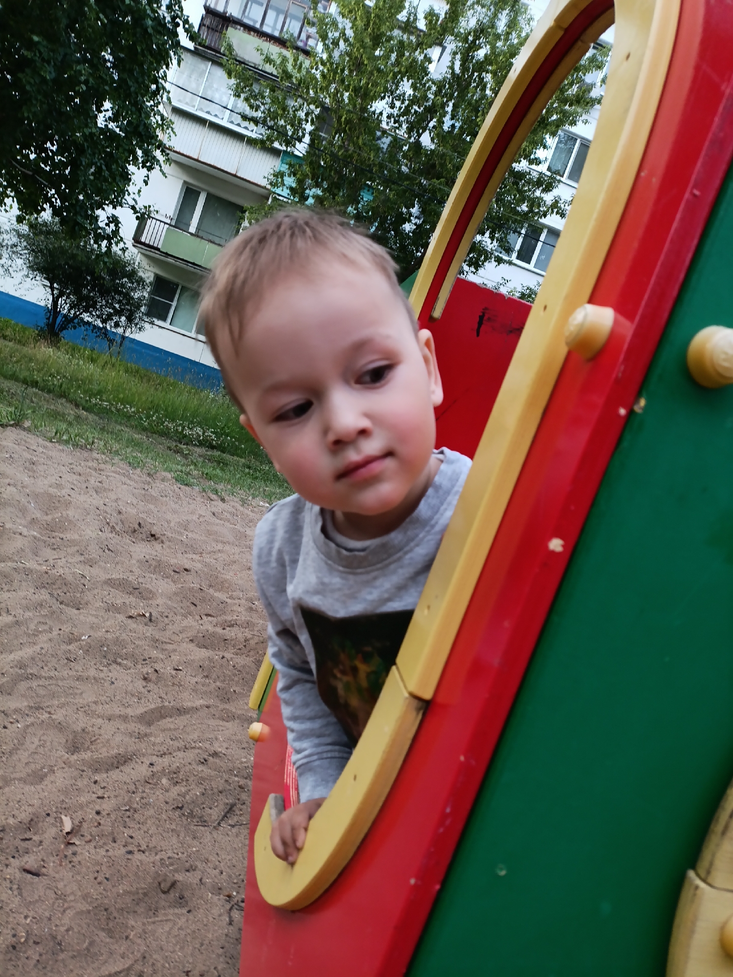 The image shows a young boy with light brown hair looking to his right. He is wearing a gray long-sleeved shirt with a colorful design on the front. The boy is partially inside a red, yellow, and green playground structure. The playground structure is made of plastic and has rounded edges and yellow knobs.
The setting is a sandy playground with patches of green grass and small white flowers visible in the background. Behind the grass, there is a tall, multi-story building with several balconies. Trees with green leaves are also present, some obscuring parts of the building. The sky is overcast, suggesting a cloudy day. The overall scene appears to be an outdoor playground in a residential area.
The boy's expression is neutral, perhaps a little pensive. He is looking downwards and to the side, away from the camera. His right hand is visible, resting on the edge of the playground structure. The image is taken from a low angle, looking up at the boy.
There is no visible text in the image. The time of day appears to be daytime, based on the lighting, and the weather is overcast. The presence of the building and playground suggests an urban or suburban environment.