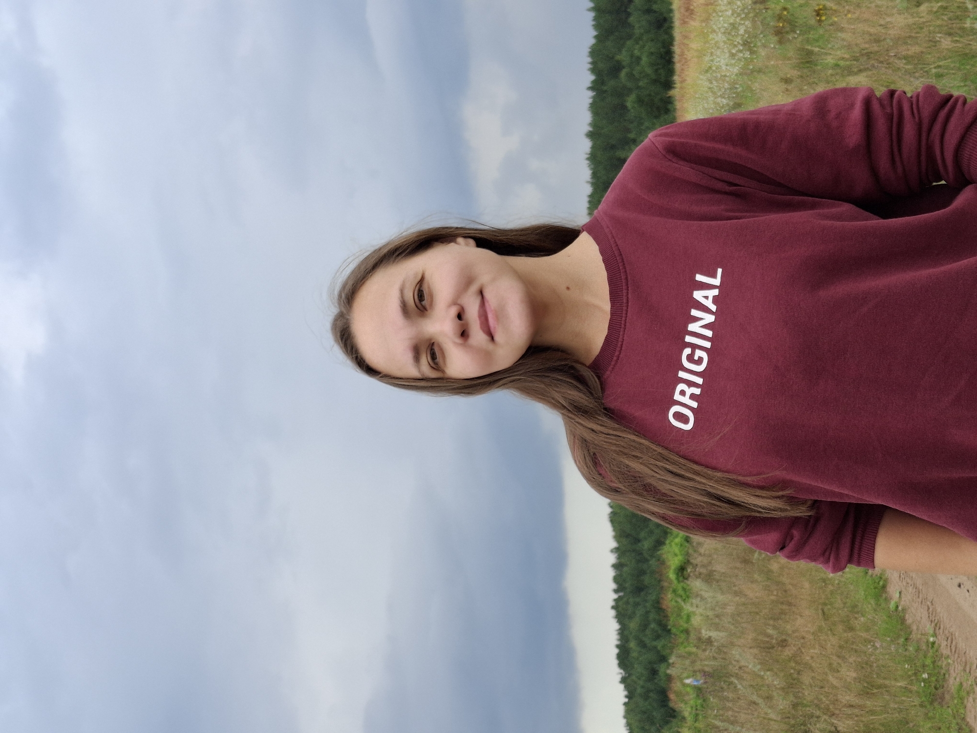 A young woman with brown hair is standing in a field with a cloudy sky overhead. She is wearing a maroon sweatshirt with the word "ORIGINAL" printed in white on the front. The field is covered in tall grass and wildflowers, and a line of trees is visible in the background. A dirt path is visible to the left of the woman. The location is the city of Plishkino, Russia.
