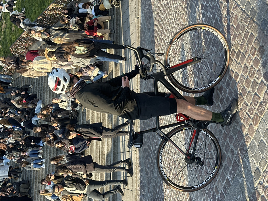 The image depicts a crowded public staircase in Montmartre, Paris, France, specifically the steps leading to the Sacré-Cœur Basilica, identified by the distinctive architecture, dense gathering of people, and love locks affixed to railings on the right.
In the foreground, a male cyclist, viewed from behind and slightly to the right, stands beside a black road bicycle with red accents. He wears a white and red MET cycling helmet, a dark neck gaiter, a black long-sleeved athletic top, black cycling shorts, olive green socks, and black "SPORT" branded cycling shoes. He holds a smartphone in his gloved right hand. The bicycle features a black frame, black saddle with a small saddle bag, a red water bottle in a cage, drop handlebars, and wheels with "Mavic" branding and tan sidewall tires. The ground is paved with cobblestones, exhibiting long shadows consistent with morning or late afternoon sun.
In the midground and background, a large assembly of approximately 50-70 individuals occupies the wide stone staircase. Most people are seated casually on the steps, facing generally towards the viewer or engaging with companions. Others are standing or walking on the steps. People are dressed in varied casual attire. On the far right, a metal railing running alongside the steps is visibly covered with numerous "love locks." Beyond the railing, a grassy slope is visible. The overall scene indicates a busy public recreational area.