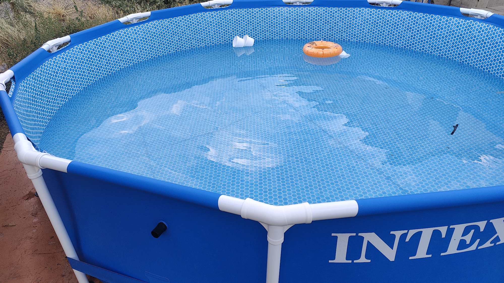 An above-ground Intex swimming pool is situated outdoors on reddish-brown, dry earth, with sparse, dry vegetation in the background. The pool's exterior is bright blue with white piping and connectors. The interior of the pool is lined with a blue hexagonal pattern and filled with clear water. Floating in the water are two white cylindrical objects and an orange ring. The "INTEX" logo is visible on the side of the pool. No people are present in the image. The scene is likely a residential backyard in Taraz, Kazakhstan, given the arid surroundings and the type of pool.