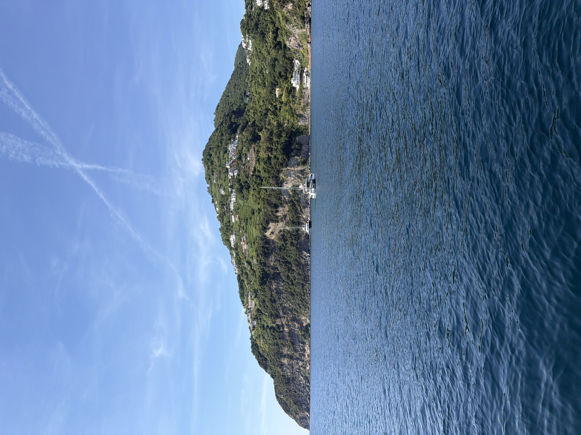 The image shows a coastline with a mountain rising from the sea, possibly in Ischia, Italy. The mountain is covered in lush greenery, and white buildings are nestled among the trees. A single white sailboat with a tall mast is anchored near the base of the mountain. The sea is calm and blue, with subtle ripples on the surface. The sky is light blue with faint contrails forming an "X" in the upper left corner. The scene is serene and typical of a Mediterranean coastal landscape.