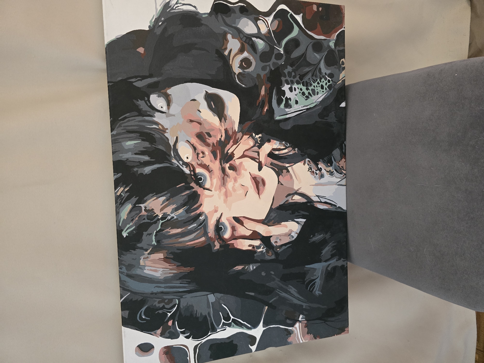 A painting of a woman's face is depicted on a canvas. The woman has dark hair and multiple eyes, some of which appear to be distorted or merging. Her skin is pale, and she is wearing dark lipstick. The style of the painting is abstract and surreal, with bold lines and a limited color palette of blacks, grays, blues, pinks, and browns. The painting is displayed on a gray surface against a neutral background. The overall impression is one of a fragmented and dreamlike portrait.