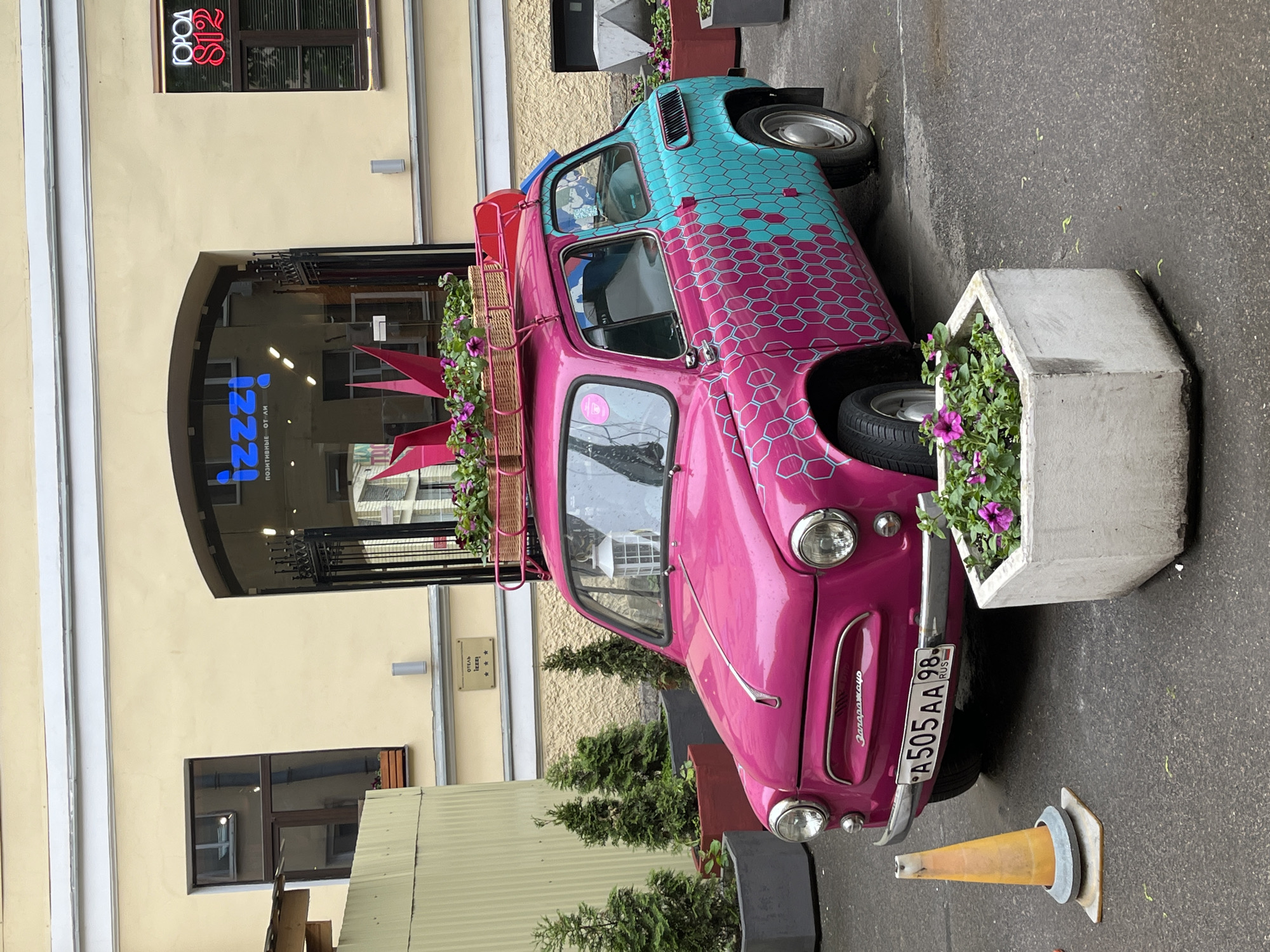 The image shows a small, vintage car, likely a ZAZ-965 Zaporozhets, decorated in a vibrant pink and blue honeycomb pattern. The car has a luggage rack on top, filled with flowers. The car is parked next to a flower planter and an orange traffic cone. The license plate reads "A505AA 98 RUS." The car is parked in front of a building with a sign that reads "izzi" and a smaller sign reading "отель izzi ***". The building facade is a light yellow color with architectural details. Another sign reads "ГОРОД S12". The location is Saint Petersburg, Russia.