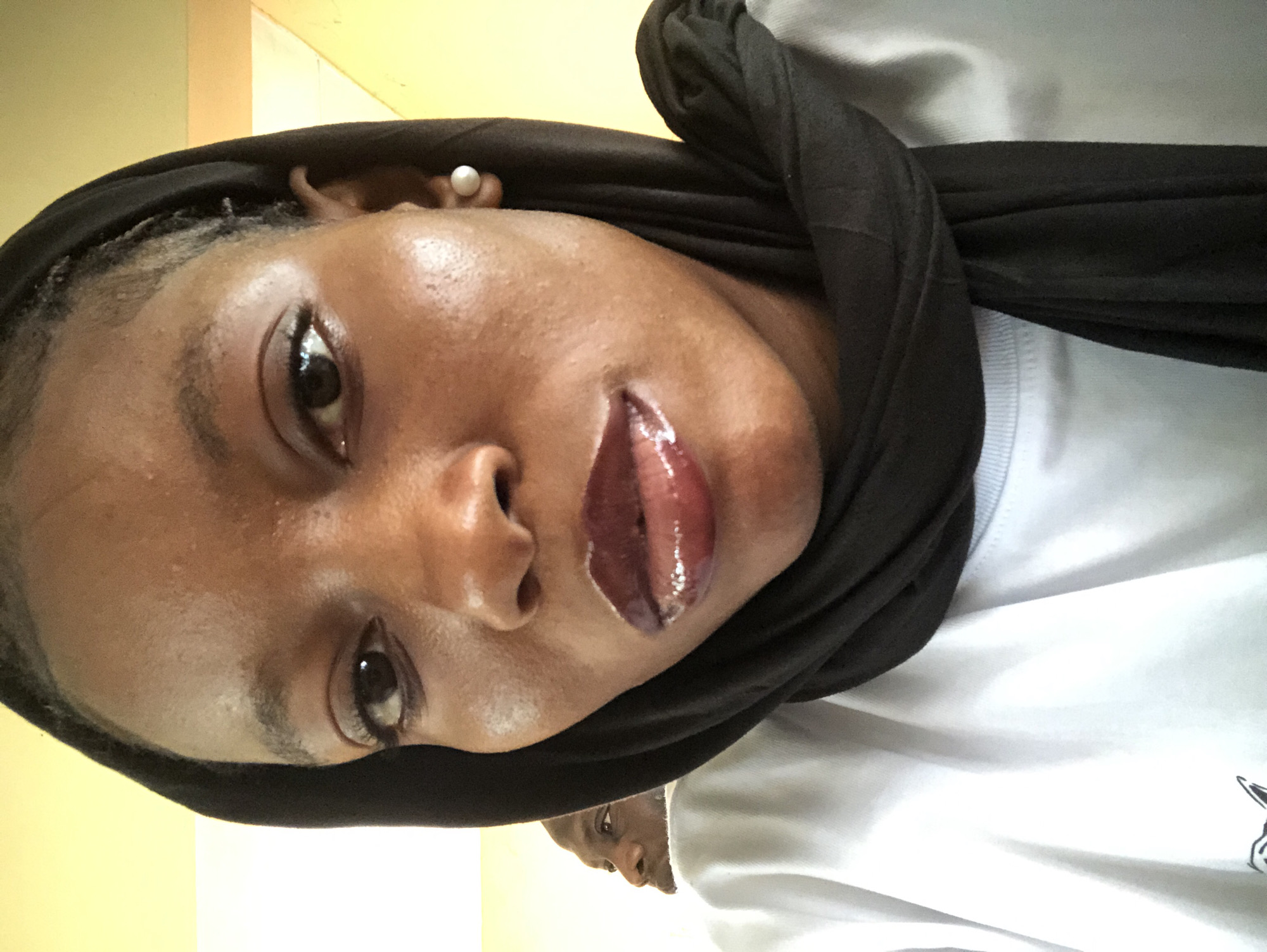 A close-up, slightly rotated image depicts a person with dark skin, wearing a black headscarf, and glossy lip color. The person appears to be wearing a white shirt. Another person's head is partially visible in the lower portion of the image. The background consists of a pale yellow wall. There is no information to indicate that the image was taken in Pindiga, Nigeria.