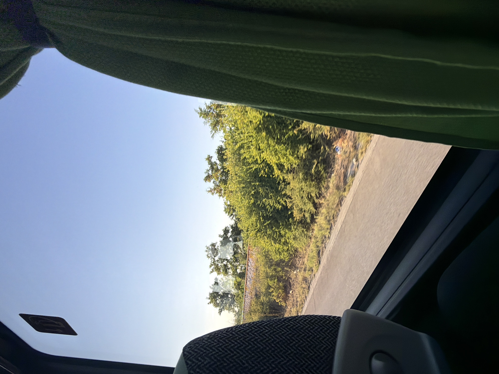 The image shows a view from inside a vehicle, likely a bus, looking out the window. The left side of the frame is occupied by the back of a seat, upholstered in a dark fabric with a patterned weave. To the right, a green curtain hangs, partially obscuring the view.
Through the window, a road runs parallel to a stretch of greenery dominated by lush trees and bushes. In the distance, beyond the vegetation, a railway track can be seen. The sky above is a clear, bright blue, suggesting a sunny day. A small, square sticker with an upward-pointing arrow is visible on the window in the upper left corner. The overall impression is of a journey through a rural or semi-rural area near Benkovski, Bulgaria, possibly on a highway adjacent to a railway line.