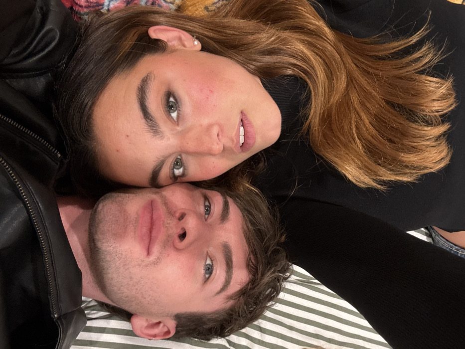 A young couple is lying down, their heads close together. The woman, positioned at the top, has long, wavy, brown hair and is wearing a black turtleneck sweater. The man, at the bottom, has short, wavy brown hair and a light stubble beard and is wearing a black leather jacket. They both are looking at the camera. The background is a bed with green and white stripes.