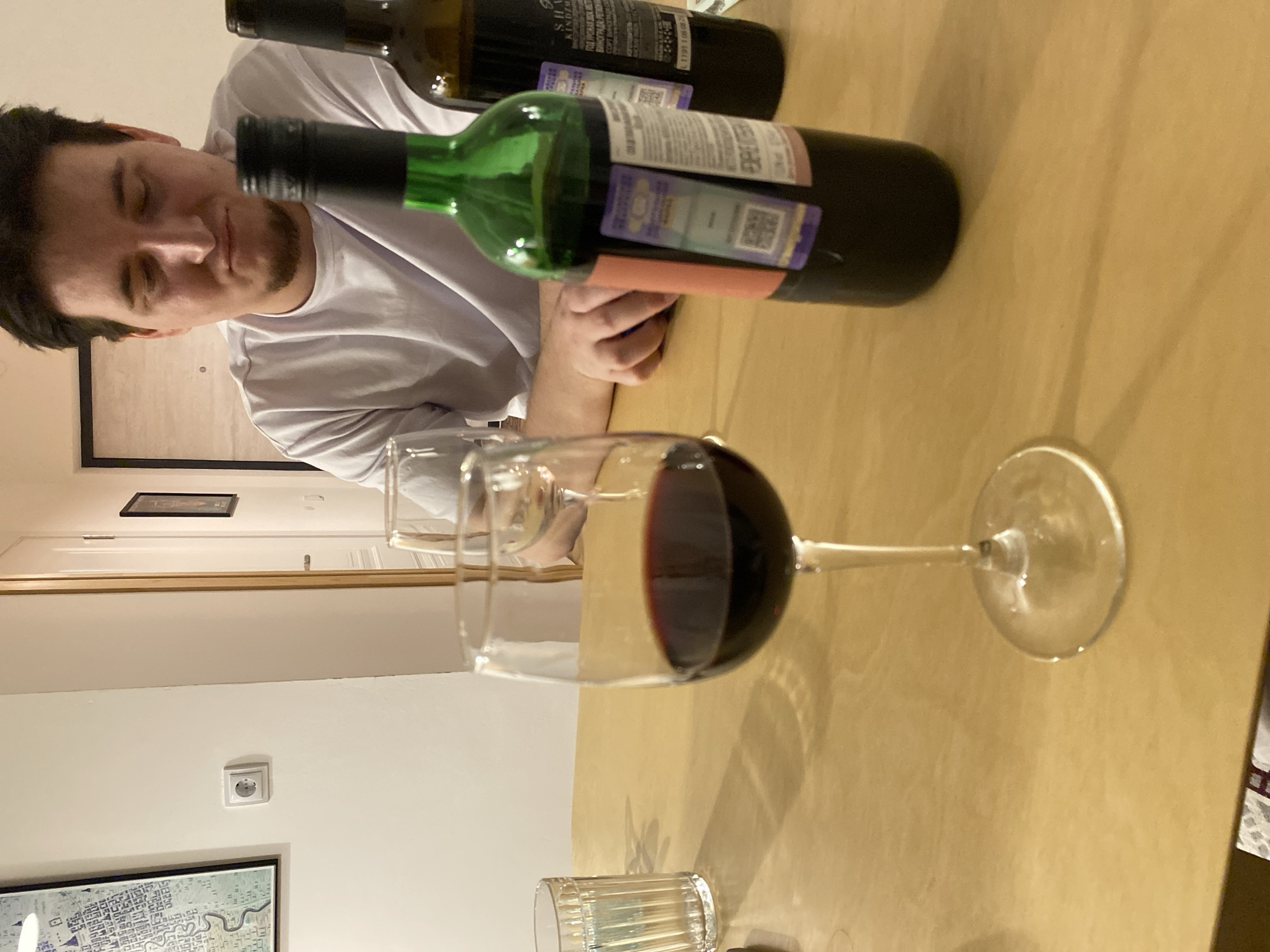 A man is seen in an indoor setting with a wine glass containing red wine and two wine bottles, one green and one dark, on a table. The man has dark hair and is wearing a white t-shirt. In the background, there's a wall with a picture frame and an electrical outlet. There is a water glass below the wine glass. The scene does not appear to be in Izhevsk, Russia, as there are no distinct indicators of the city present.
