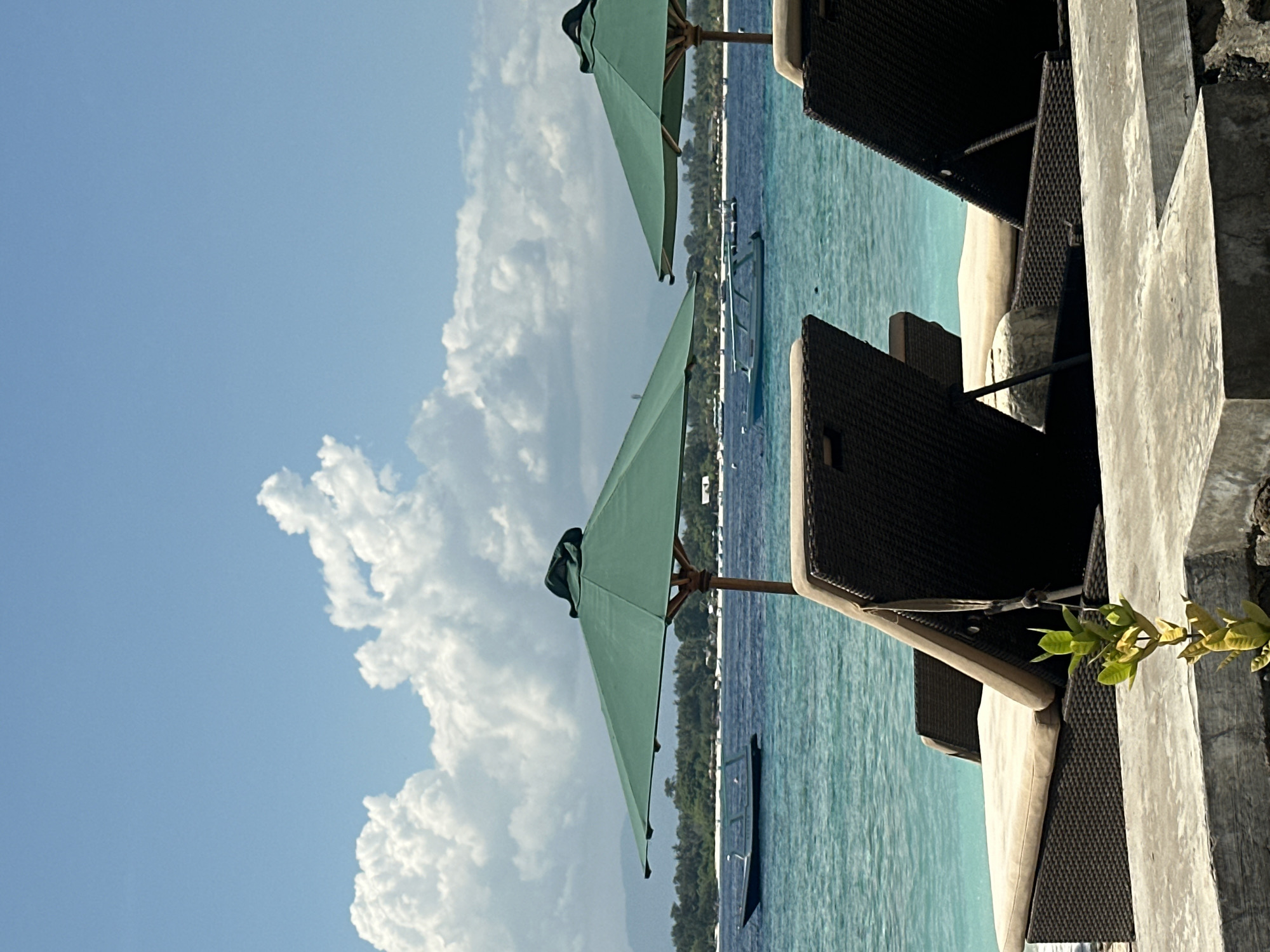 The image shows a coastal scene, likely taken in an outdoor seating area of a resort or hotel in Unknown, Indonesia. Two green umbrellas provide shade over dark brown wicker chairs, with an ocean view and blue sky filled with puffy white clouds. The turquoise water is calm, and a boat is visible in the distance. A textured, light gray wall borders the seating area, with some green plants growing near it.