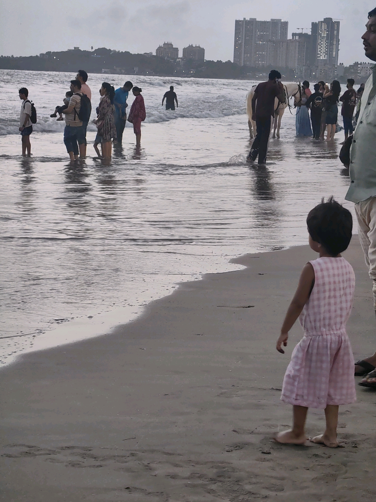 The image depicts a crowded beach scene in Mumbai, India, under an overcast sky. In the foreground, a young child in a pink checkered dress stands on the wet sand, facing away from the camera and towards the ocean. Behind the child, a group of people are wading in the shallow water, some with children. Further back, a white horse is visible in the water with a person leading it. In the background, a cityscape with several tall buildings is visible, indicating an urban coastal environment. The water is moderately rough with visible waves breaking on the shore.