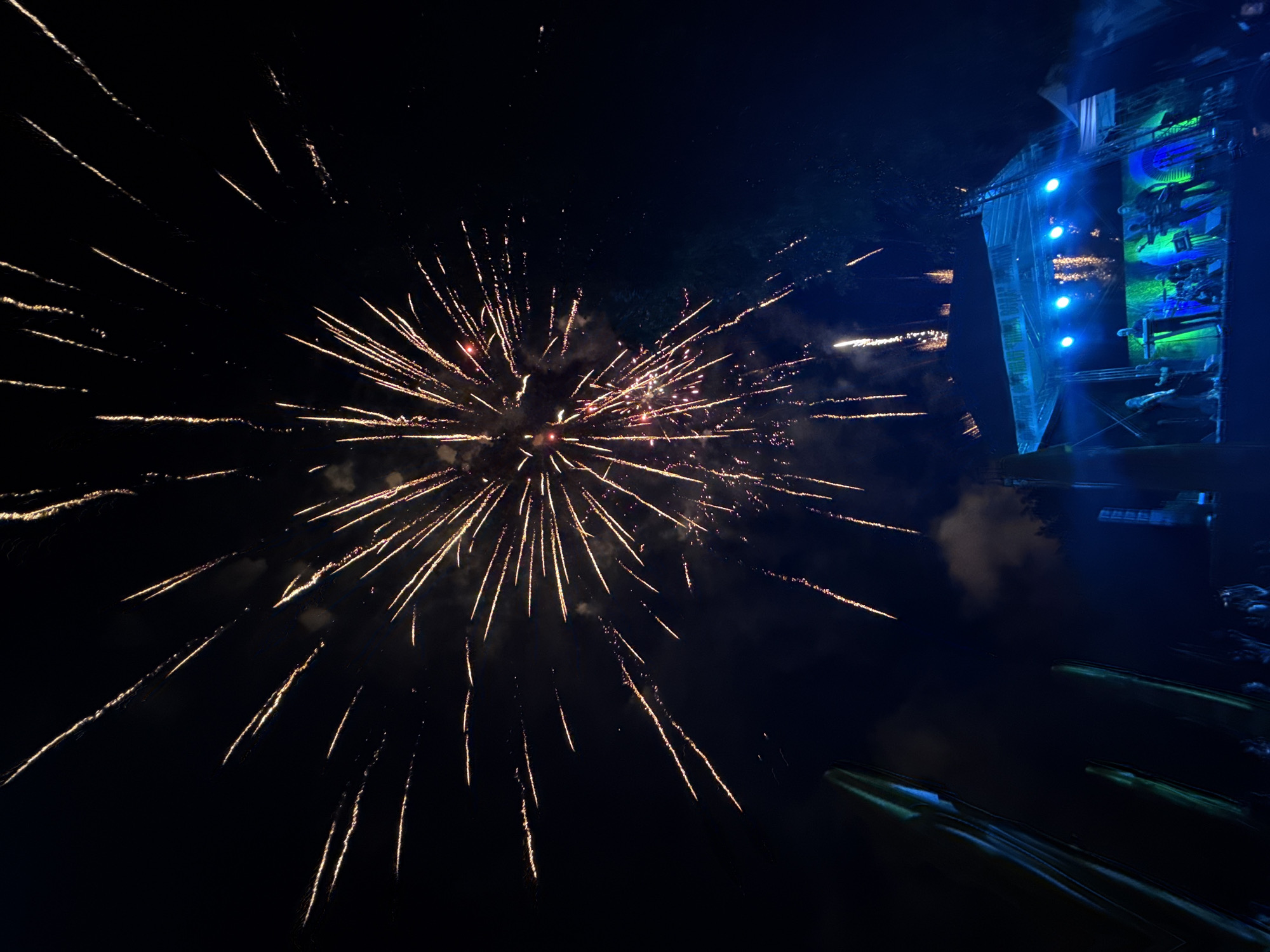 At night, in the city of Gara Oreshets, Bulgaria, fireworks are exploding in the dark sky, with a large, bright burst dominating the upper half of the image. In the lower portion, a stage with a band performing is illuminated by blue and green lights, with silhouettes of people visible. In the foreground on the left, there are tall, abstract green structures, possibly part of the stage lighting or decorations.