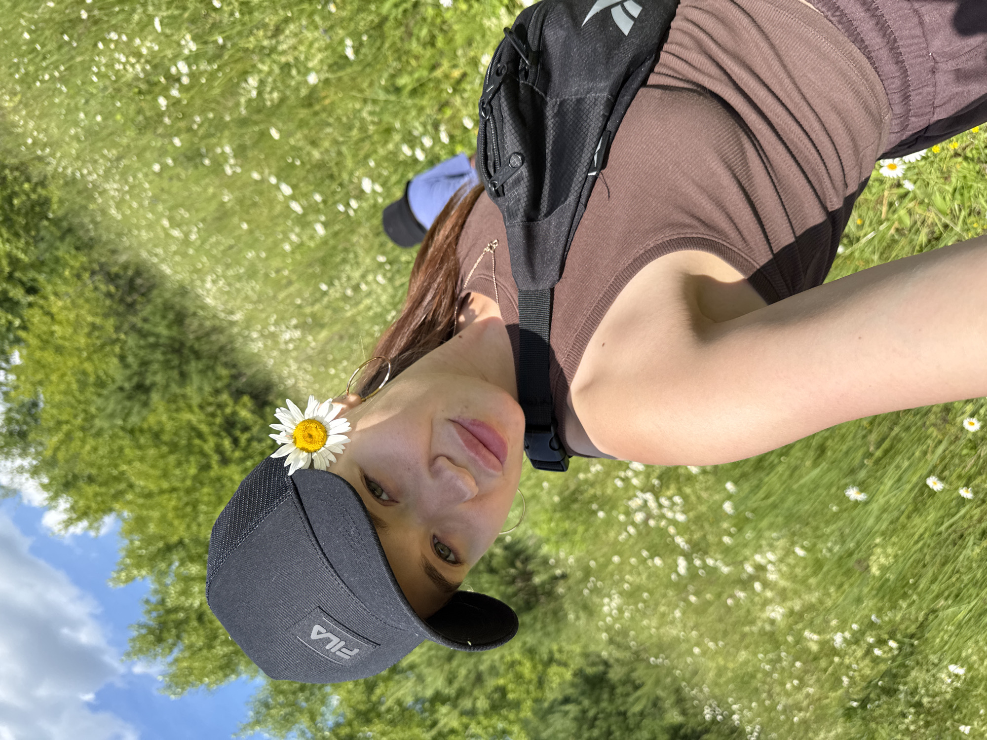 This is a portrait of a young woman lying in a grassy field speckled with white wildflowers. She is wearing a brown ribbed tank top, a black fanny pack with a silver logo, and a black baseball cap with "FILA" printed on it. A daisy is tucked behind her ear. The woman has green eyes and is smiling at the camera. The background consists of lush green grass and trees, with a blue sky and white clouds visible. The lighting suggests it is a sunny day, likely in the late morning or afternoon. There is no explicit indication of the activity or event, but the pose and setting suggest a relaxed outdoor scene. The location is identified as Гаревая, Russia.