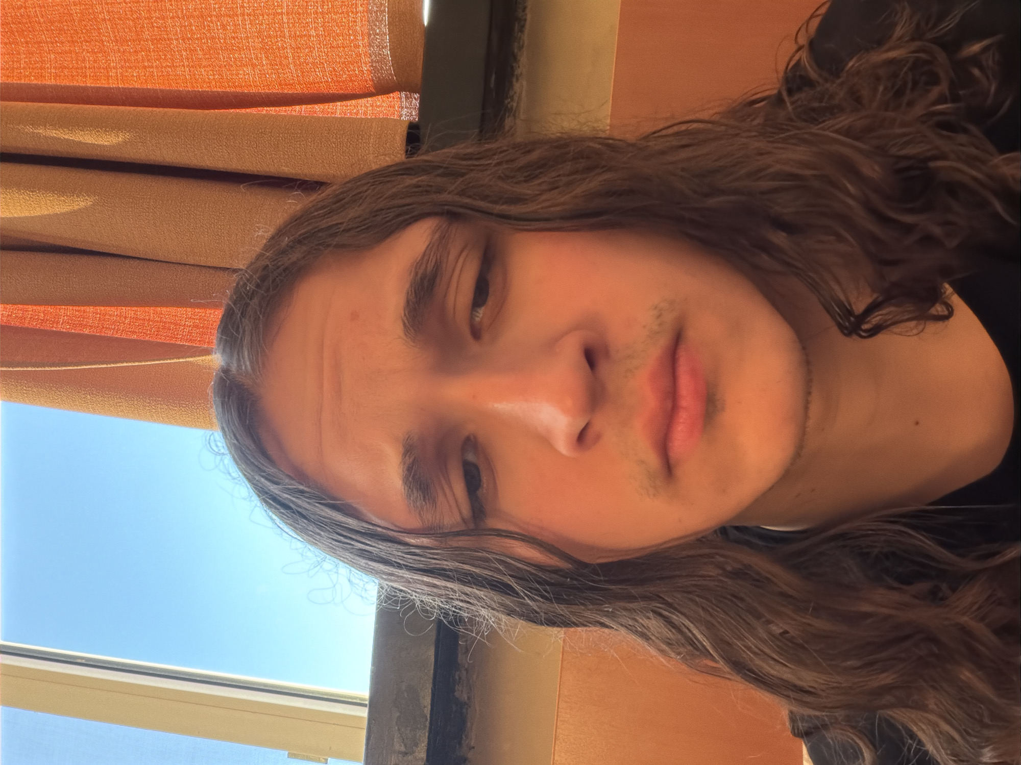 A close-up, rotated portrait captures a young individual with long, wavy dark brown hair, looking directly at the viewer with a neutral or slightly pensive expression. The person's left eye (viewer's right) appears slightly narrower than their right. Faint stubble is visible on the chin and upper lip.
The setting is indoors, with bright natural daylight illuminating the scene. A clear blue sky is visible through a window in the bottom left corner of the original image, indicating it is daytime. Orange and brown blinds or curtains serve as the background on the top and right sides of the frame. The subject is wearing a black garment, parts of which are visible on the right side of the image. The hair is somewhat dishevelled, framing the face. There is no visible text in the image. The location context provided is Plovdiv, Bulgaria.