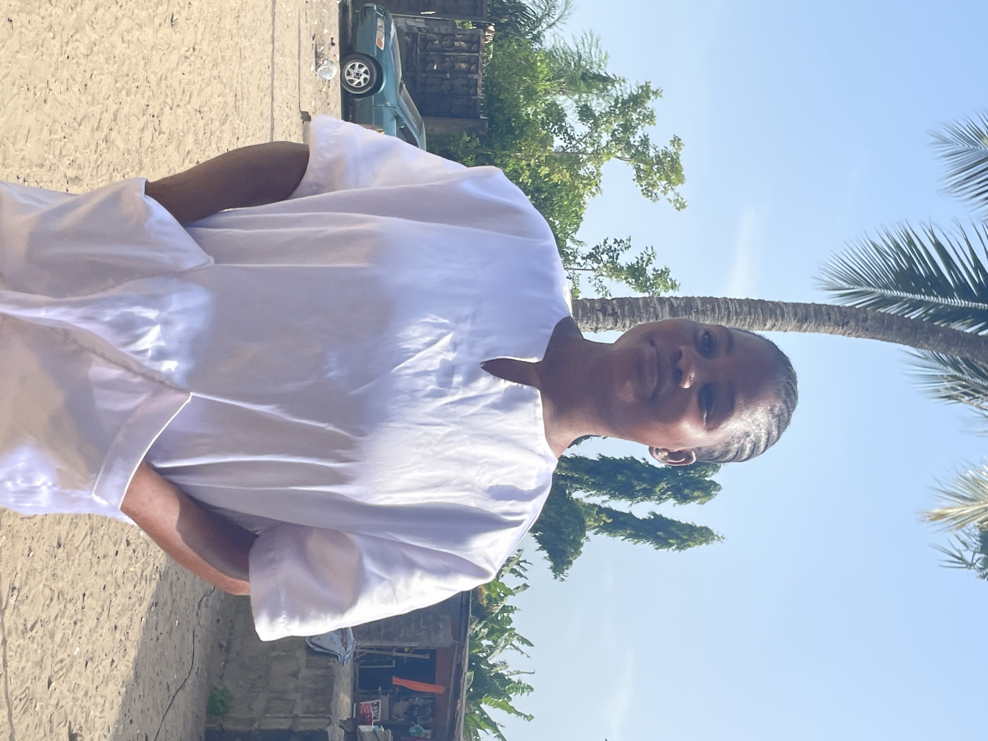 A person wearing a white garment stands outdoors. Behind the person are trees and foliage, with a blue sky visible above. In the background, a section of a building is visible, along with what appears to be a parked car. Based on the information given, the location is Badagry, Nigeria.