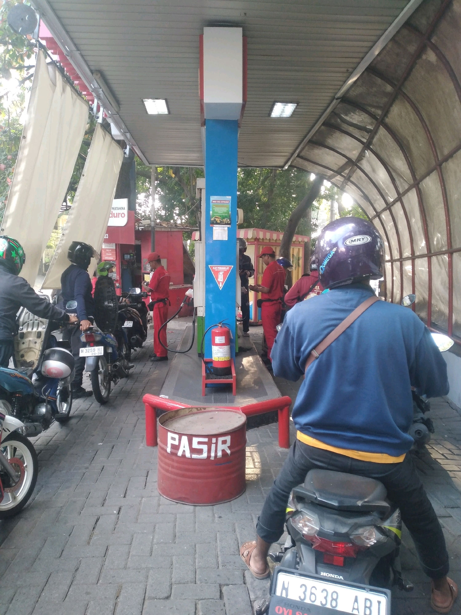 The image depicts a scene at a Pertamina gas station, possibly located in the city of Malang, Indonesia, as suggested by the license plate visible on a motorcycle. The gas station has a covered canopy with a corrugated metal roof, supported by a blue central pillar. Several motorcycles are parked, and people are present, engaged in activities related to refueling.
In the foreground, a person wearing a blue long-sleeved shirt, black pants with a yellow waistband, and a purple helmet is seated on a motorcycle. They are facing away from the camera. The motorcycle has a license plate that reads "N 3638 ABI". To the left of this motorcycle, another motorcycle is partially visible, with its front wheel and fender showing.
Further back, a gas pump is visible, with a red hose extending from it. A person in a red uniform, likely a gas station attendant, is standing near the pump, interacting with a customer or preparing to refuel. Several other individuals are also seen around the motorcycles and the service area, some wearing helmets.
A red metal barrel with the word "PASIR" (Indonesian for sand) written in white letters on its side is placed on the ground, to the right of the foreground motorcycle and in front of the gas pump. A red fire extinguisher is mounted on a stand near the gas pump.
The ground is paved with interlocking bricks. In the background, trees and foliage are visible, suggesting an outdoor setting. The overall atmosphere appears to be a typical busy moment at a gas station.