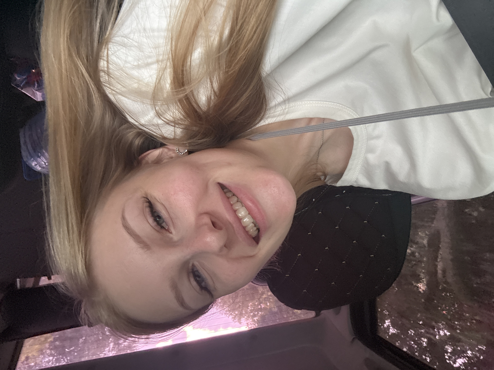 A young woman with blonde hair smiles into the camera from inside a vehicle. The background appears to be a wooded area seen through the car window, tinted with a purple hue. The woman is wearing a white t-shirt, and a grey drawstring is visible across her chest. The interior of the car is dimly lit, with a dark-colored headrest visible behind her. The image is a selfie, suggesting the woman is taking the picture herself. The context of being in the city of Perm, Russia, is not discernible from the visual information alone.