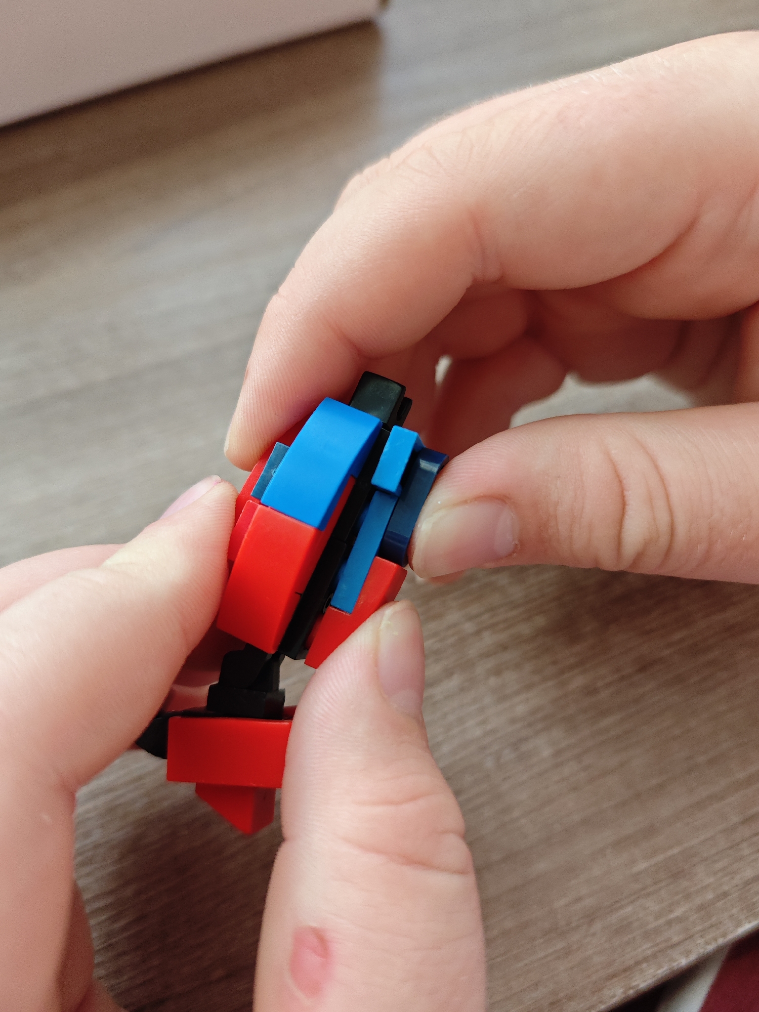 A close-up shot shows two hands, possibly belonging to a child, assembling a small construction toy made of red, blue, and black interlocking plastic bricks. The toy is held at an angle, with one hand supporting the base and the other finger precisely connecting a blue brick to the red and black structure. The background is a blurred wooden surface, likely a table, in an indoor setting. The lighting is soft, suggesting natural light. There is no discernible location in Марковский, Russia, or any other city.