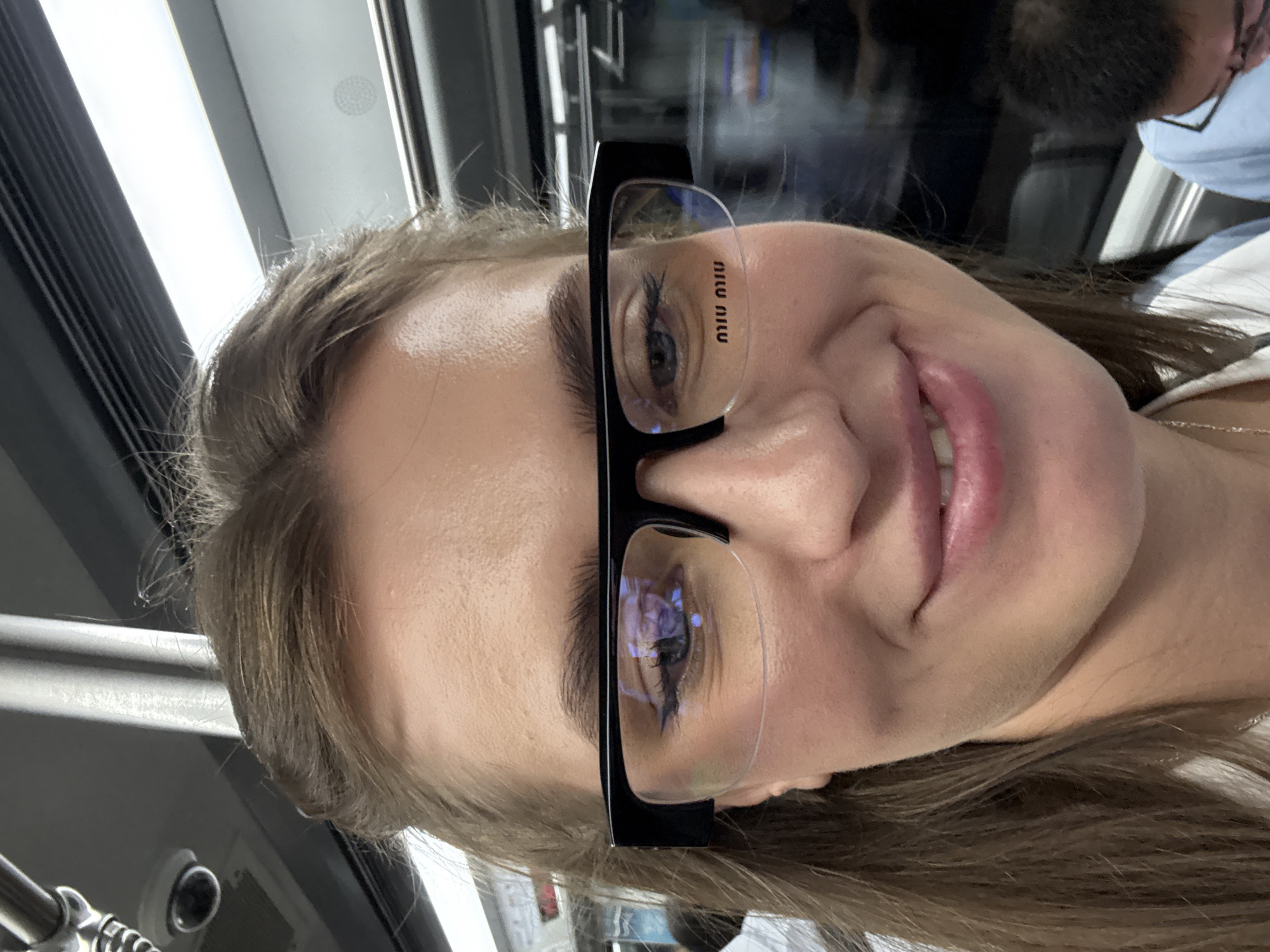 A close-up selfie of a young woman wearing black-rimmed glasses with "MIU MIU" written on the lenses. She has her hair pulled back and is smiling. The background appears to be the interior of a train, with a metal pole and a security camera visible. The image is taken at eye level. The location is Haizhu District, China.