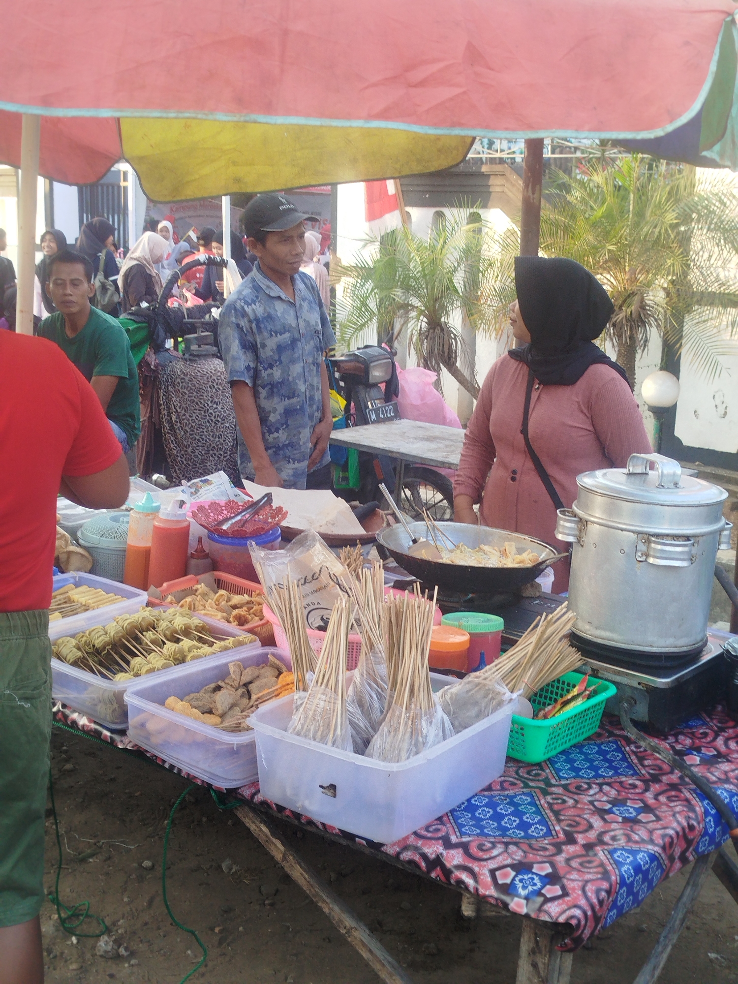The image captures an outdoor food stall in Burneh, Indonesia, under a red and yellow awning. A man wearing a blue patterned shirt and a black baseball cap stands behind a table laden with food items. To his right, a woman in a pink long-sleeved shirt and black hijab is also behind the table, attending to a large metal pot. In front of them are various containers filled with food, including skewers of what appear to be fried snacks and other processed food items. Several bottles of sauces and condiments are visible, along with a wok containing food being cooked. Sticks and other cooking utensils are also present. In the background, more people are milling around, some under the awning, and palm trees and a building are visible. The stall appears to be situated on a dirt ground.