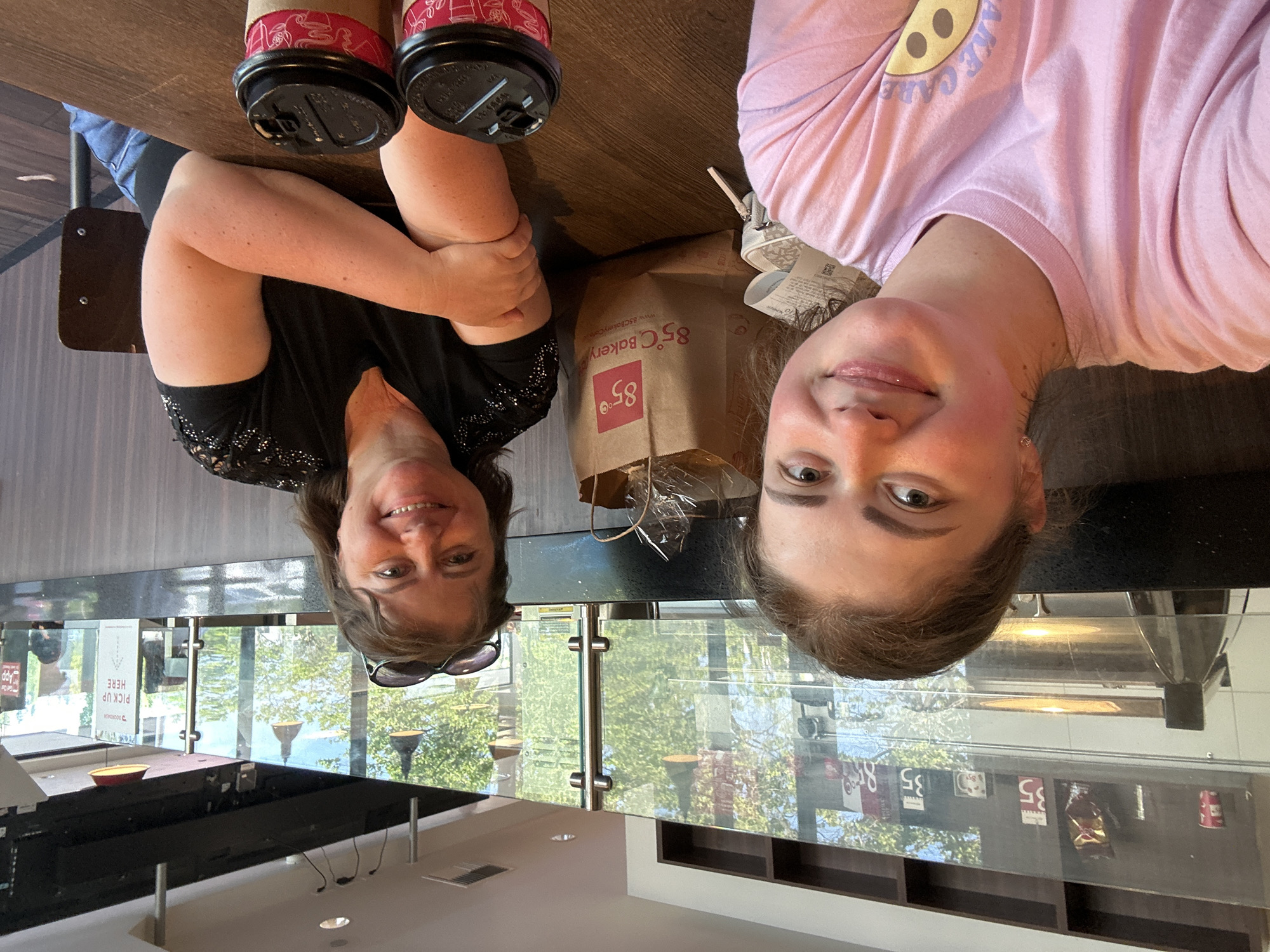The image depicts two women sitting at a wooden table in what appears to be a cafe or bakery, likely at an "85°C Bakery Cafe" given the prominent branding on a paper bag between them. The cafe features a modern aesthetic with a glass partition separating the seating area from the kitchen or service counter, through which windows reveal trees and sunlight. The woman on the left, closer to the camera, is wearing a pink t-shirt and has her hair pulled back. The woman on the right, wearing a black shirt, is also smiling and has sunglasses on her head. On the table, two coffee cups are visible, along with the bakery bag and a small white pouch. The environment suggests a casual, daytime setting. The specific location in Lynnwood, United States of America, is inferred from the presence of an 85°C Bakery Cafe, which has a branch in that city.