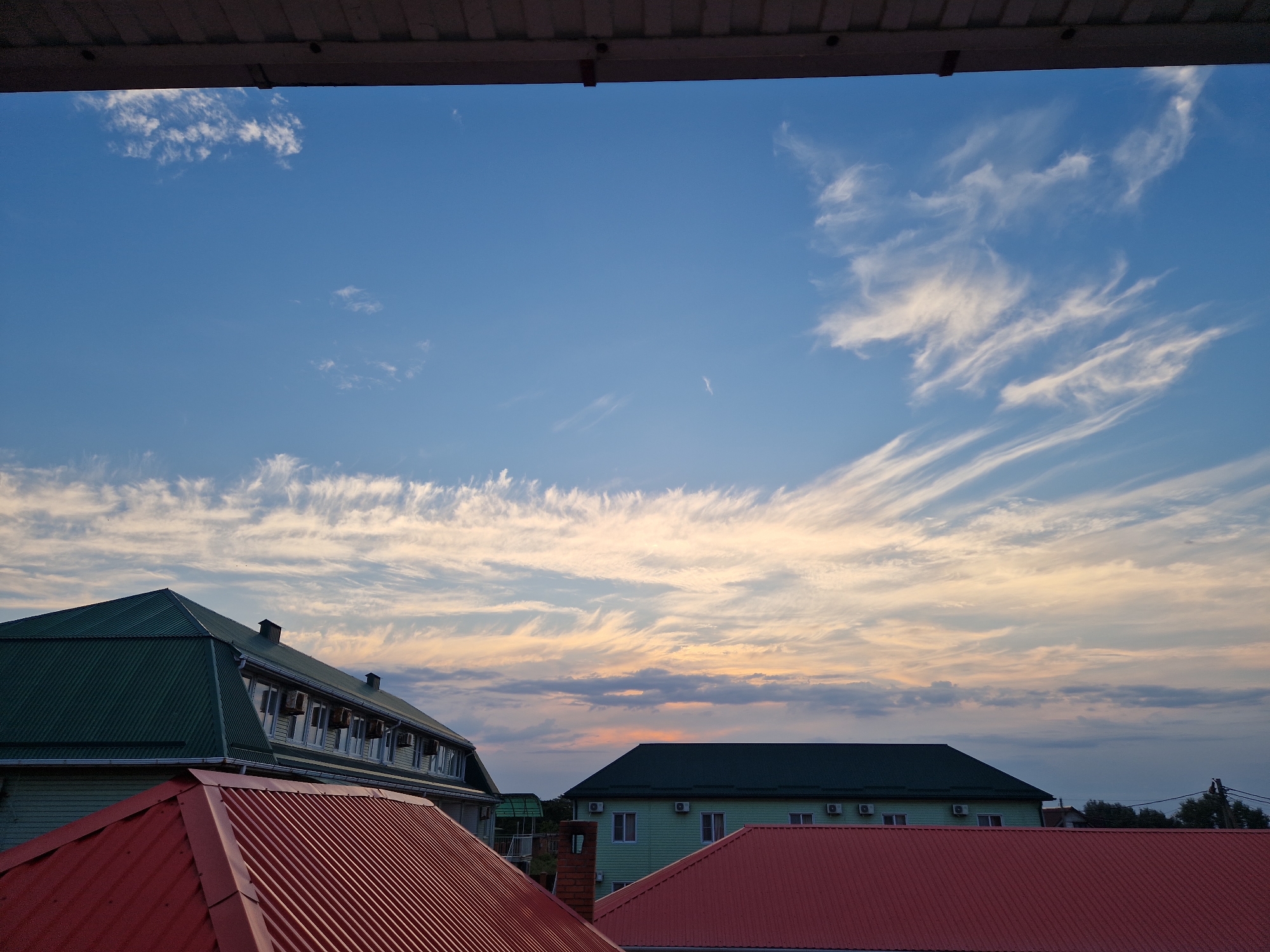 The image shows a view of several buildings from a rooftop in Peresyp, Russia, during what appears to be sunset or sunrise. In the foreground, a red corrugated metal roof dominates the lower portion of the frame. Behind it, a green corrugated metal roof of another building is visible, with multiple windows and air conditioning units. Further in the background, a light green building with similar windows and air conditioning units can be seen. The sky above is a clear blue, with scattered wispy white clouds tinged with orange and pink hues, indicating the time of day. The scene is static, with no people or actions depicted. The overall impression is one of a residential or small commercial area under a dramatic sky.