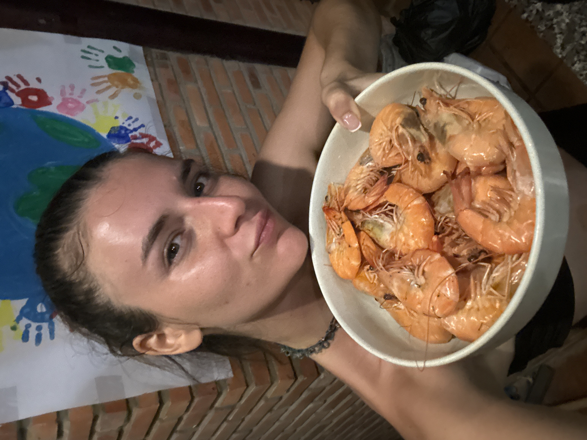 A young adult female is depicted holding a white ceramic bowl filled with approximately 15-20 whole, cooked, orange-pink shrimp, complete with heads and shells. Her dark hair is pulled back, and she wears a dark garment, featuring a subtle beaded choker and a French manicure on her visible right hand. She looks directly at the camera with a slight, confident smile. The scene is located in Mũi Né, Vietnam. The background consists of a reddish-brown brick wall. Affixed to the upper part of the wall is a white poster displaying a large blue and green depiction (possibly of the Earth) surrounded by multiple colorful handprints (red, pink, yellow, blue, orange, green). A dark vertical structural beam is visible on the far right of the frame, and a section of a patterned rug and a black plastic bag are visible on the dark floor in the lower right.