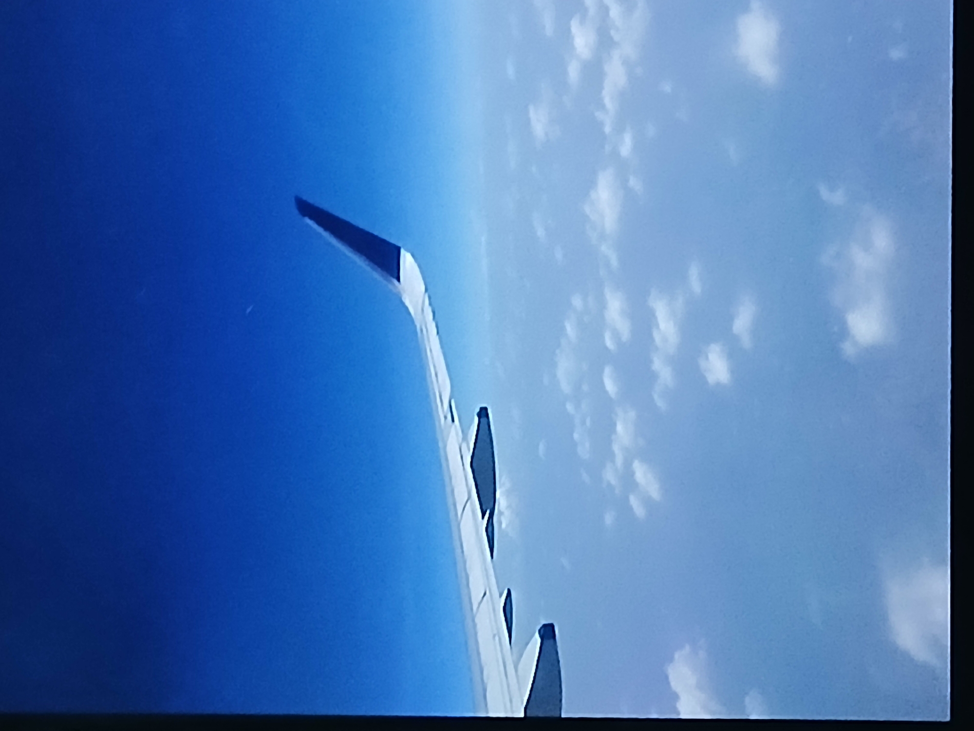 The image shows an airplane wing in flight, with a view of the sky and clouds from the airplane window. The sky on one side of the wing is a clear, deep blue, while the other side contains scattered white clouds. The image does not contain information or details about the city of Bengaluru, India. There are no people visible in the image.
