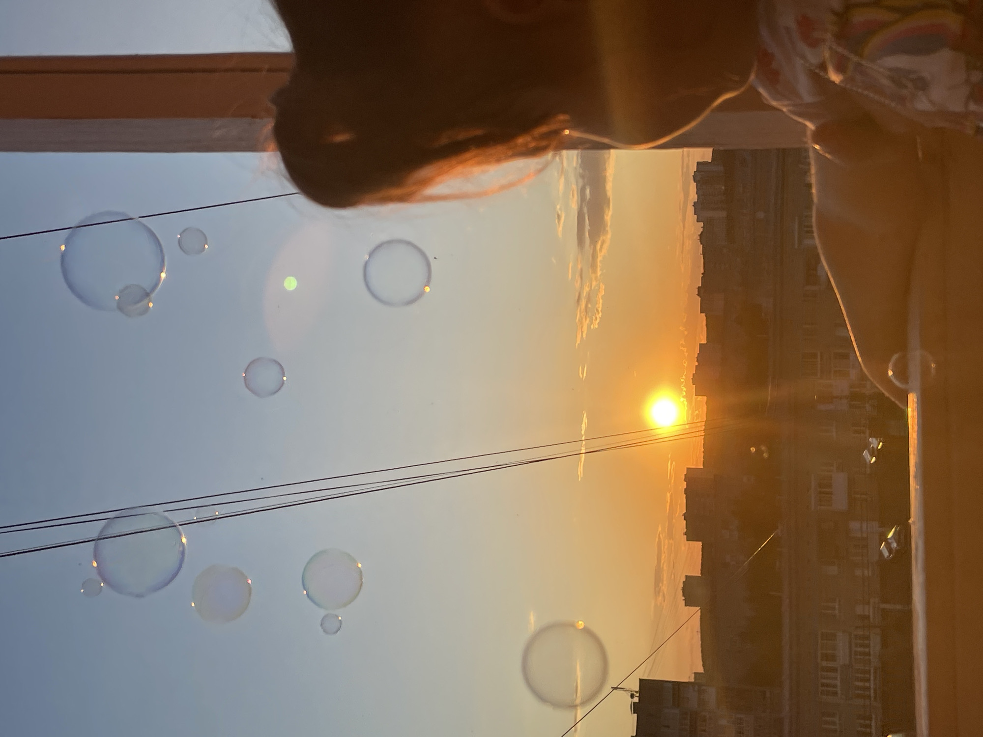 The image depicts an urban sunset scene in Izhevsk, Russia, viewed from an elevated perspective. The sky transitions from a brilliant orange-yellow at the horizon, where the circular, intense sun is prominently visible, to a lighter blue-grey in the upper atmosphere. Multiple spherical, iridescent bubbles are suspended in the air, reflecting the warm light. Several thin, dark, parallel utility cables diagonally cross the upper-right portion of the frame. A distinct bright green circular optical artifact is present in the upper-mid sky. In the immediate foreground on the right, the back of a child's head and upper torso are visible in silhouette, with strands of hair illuminated by the setting sun. The child is oriented towards the sunset, with an arm resting on a horizontal, sun-drenched surface, likely a balcony railing or window sill, upon which two small, reflective droplets or bubbles are visible. A faint rainbow pattern is discernible on a piece of the child's clothing on the lower right. The midground features a densely built cityscape comprising numerous multi-story residential buildings, silhouetted against the bright horizon, interspersed with dark vegetation. A thick, rectangular, vertical structural element, consistent with a frame or pillar, extends along the far right edge of the image.