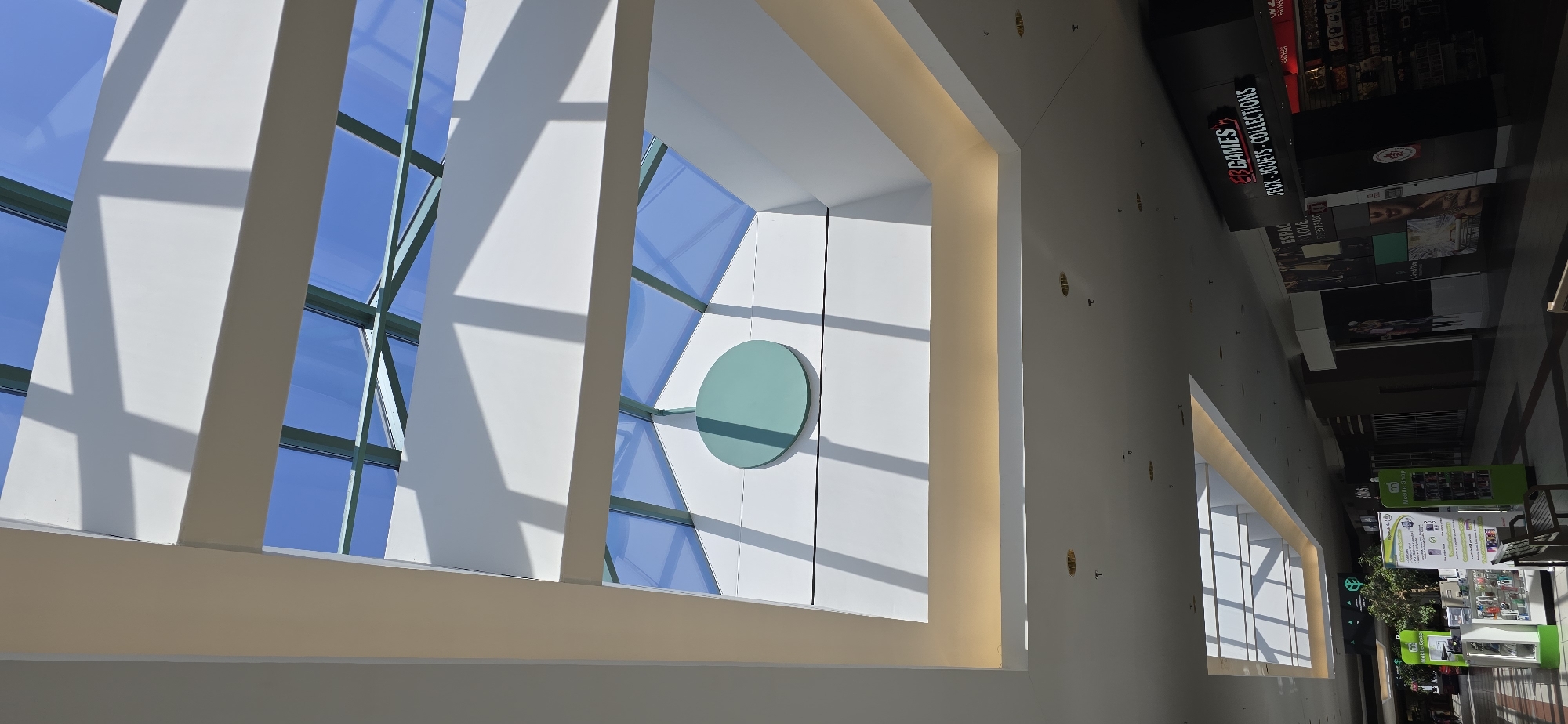 The image depicts the interior of a modern shopping mall with a prominent skylight. The skylight, constructed with glass panels and a metal frame, allows natural light to illuminate the space, casting geometric shadows on the white ceiling and walls. A large, circular, teal-colored decoration is visible on the upper part of the wall beneath the skylight. The lower portion of the image shows a mall concourse with various shops. One shop, labeled "GAMES" and "JEUX - JOUETS - COLLECTIONS," is clearly visible on the right. To the left, other retail displays and entrances are present, along with seating areas. The overall environment suggests a well-lit and contemporary commercial space. This mall is identified as being located in Victoriaville, Canada.