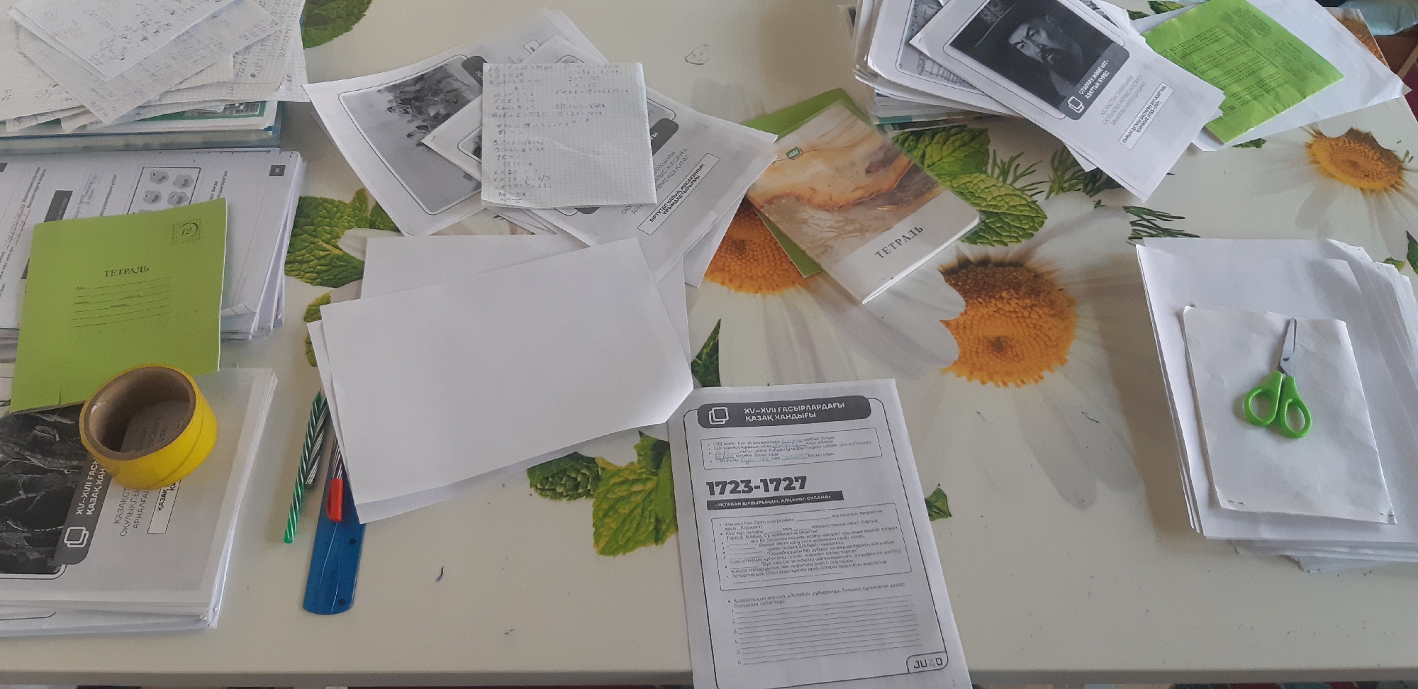 The image shows a close-up, overhead view of a table covered with various papers, notebooks, and writing utensils. A green notebook with the word "ТЕТРАДЬ" (notebook) is visible on the left side, next to a roll of yellow tape. To the right of the green notebook, a stack of white papers is partially obscured by another notebook with a patterned cover. Further to the right, a small pair of green scissors rests on a stack of white paper. In the lower right corner, a document with text in Kazakh is open, displaying a title that appears to be "XV-XVII ҒАСЫРЛАРДАҒЫ ҚАЗАҚ ХАНДЫҒЫ" (Kazakh Khanate in the XV-XVII Centuries). Pens and a ruler are also scattered on the table. The background is a table surface with a repeating pattern of daisies. There are no people or animals present in the image. The scene appears to be a workspace, possibly a desk, cluttered with educational or administrative materials.