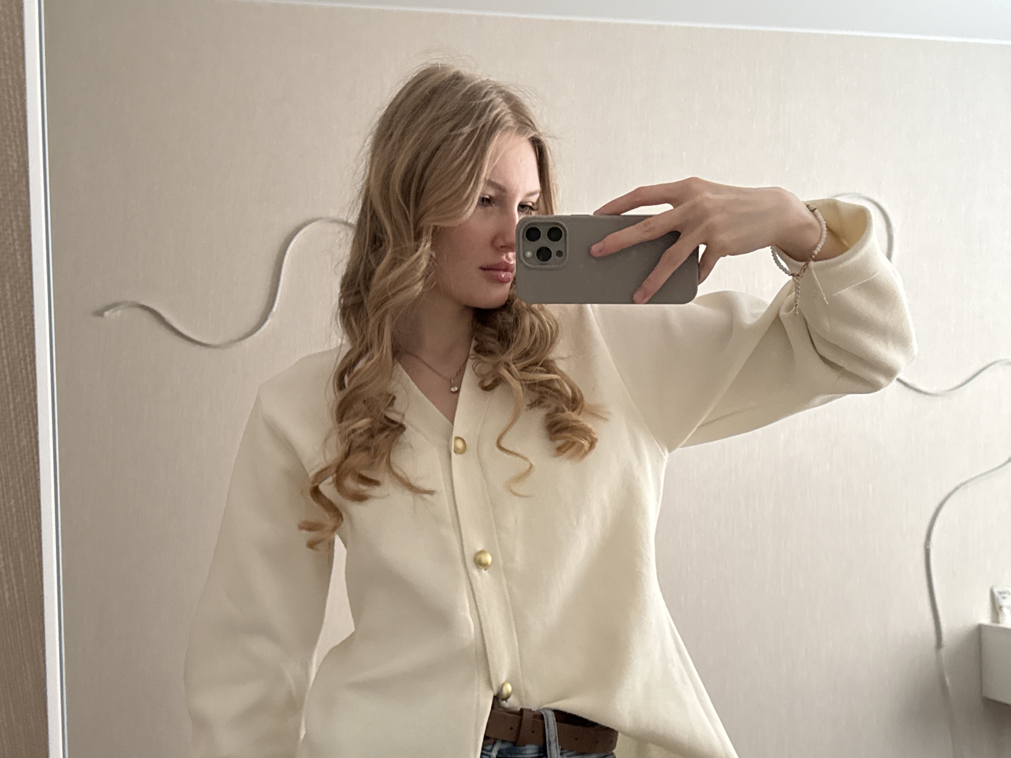 A young woman with wavy blonde hair is taking a selfie in front of a mirror. She is holding a gray smartphone in her right hand, partially obscuring her face. The woman is wearing a light cream-colored cardigan with gold buttons, a brown leather belt, and a pearl bracelet. The background consists of a light-colored wall with decorative molding. A partial view of a white box and cable is visible on the lower right. The image location may be Perm, Russia.
