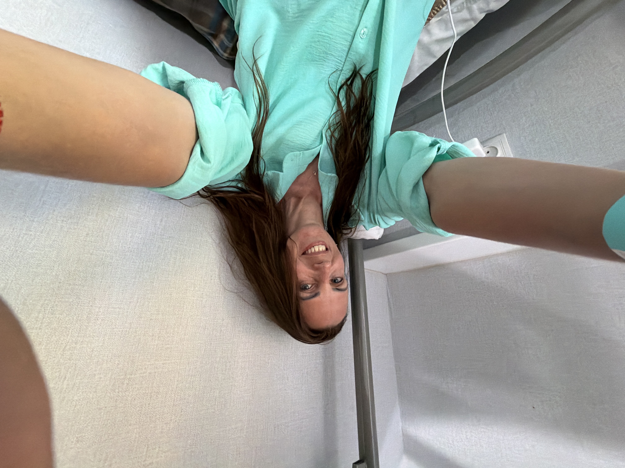 A woman with long, brown hair, wearing a turquoise blouse, smiles at the camera. She is in a room with textured walls, a metal pole, and a power outlet visible. Her arms are extended forward, suggesting she is taking a selfie. The background indicates an indoor setting, possibly a dormitory or a small living space. There are no specific landmarks visible in this image that would allow for precise location identification within Birobidzhan, Russia.