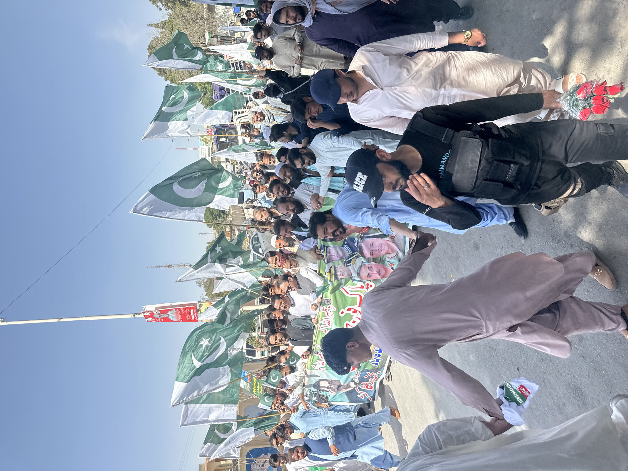 A daytime image shows a crowd of people gathered outside in Quetta City Tehsil, Pakistan. The people are carrying flags of Pakistan and a banner with images of people on it. A man in a black vest with "COMMANDO" printed on it is standing near the front. Some people are holding bouquets of red flowers. One person is reaching out to hand another a package labeled "INDOMITABI."