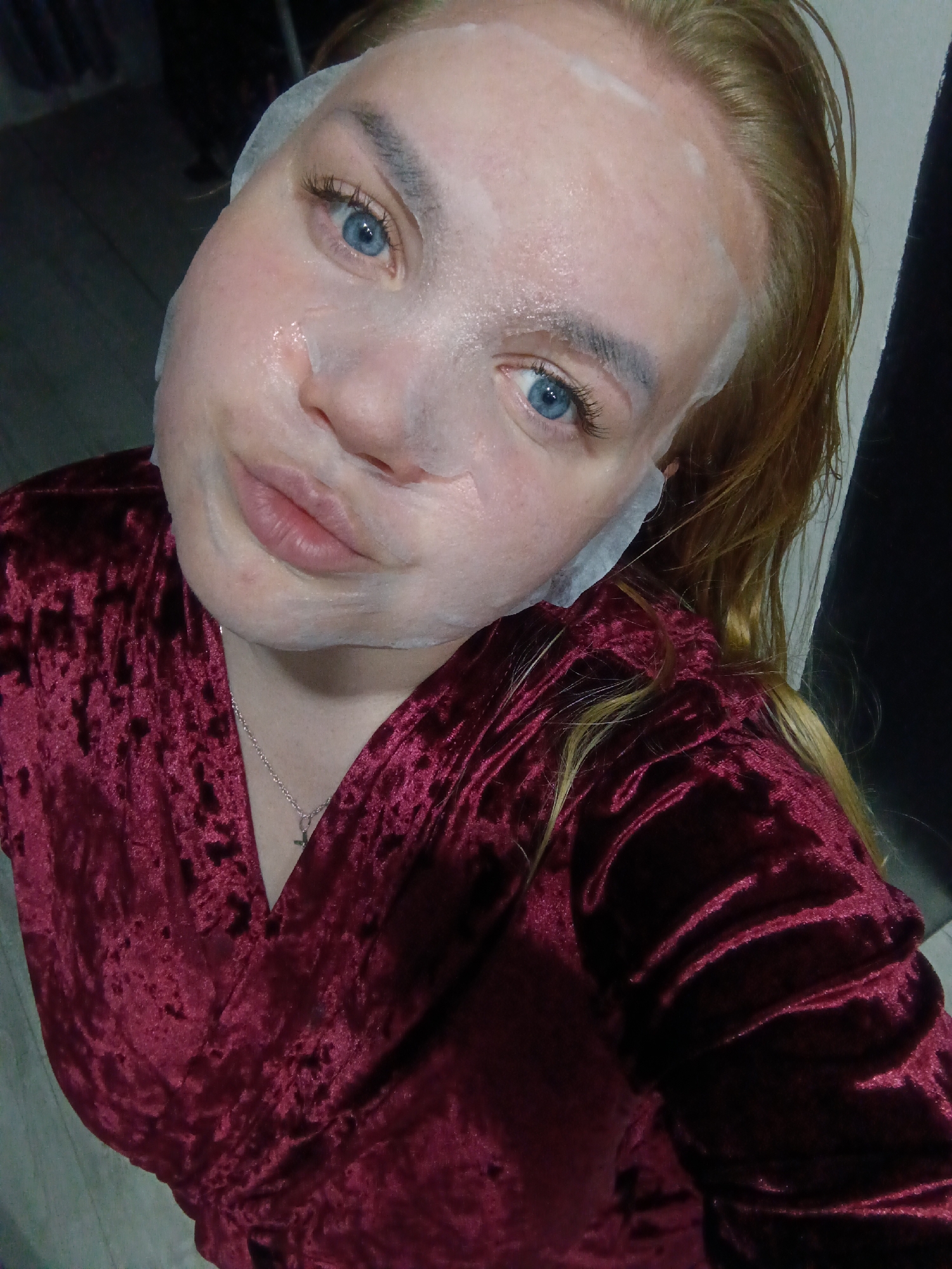 A woman with light skin and blue eyes is wearing a white sheet mask on her face. She is wearing a maroon velvet top. A silver necklace with a small cross pendant is visible. Part of the room she is in can be seen: a light grey floor and a dark wall. It is not possible to determine if the woman is in Фоки, Russia, from this image.