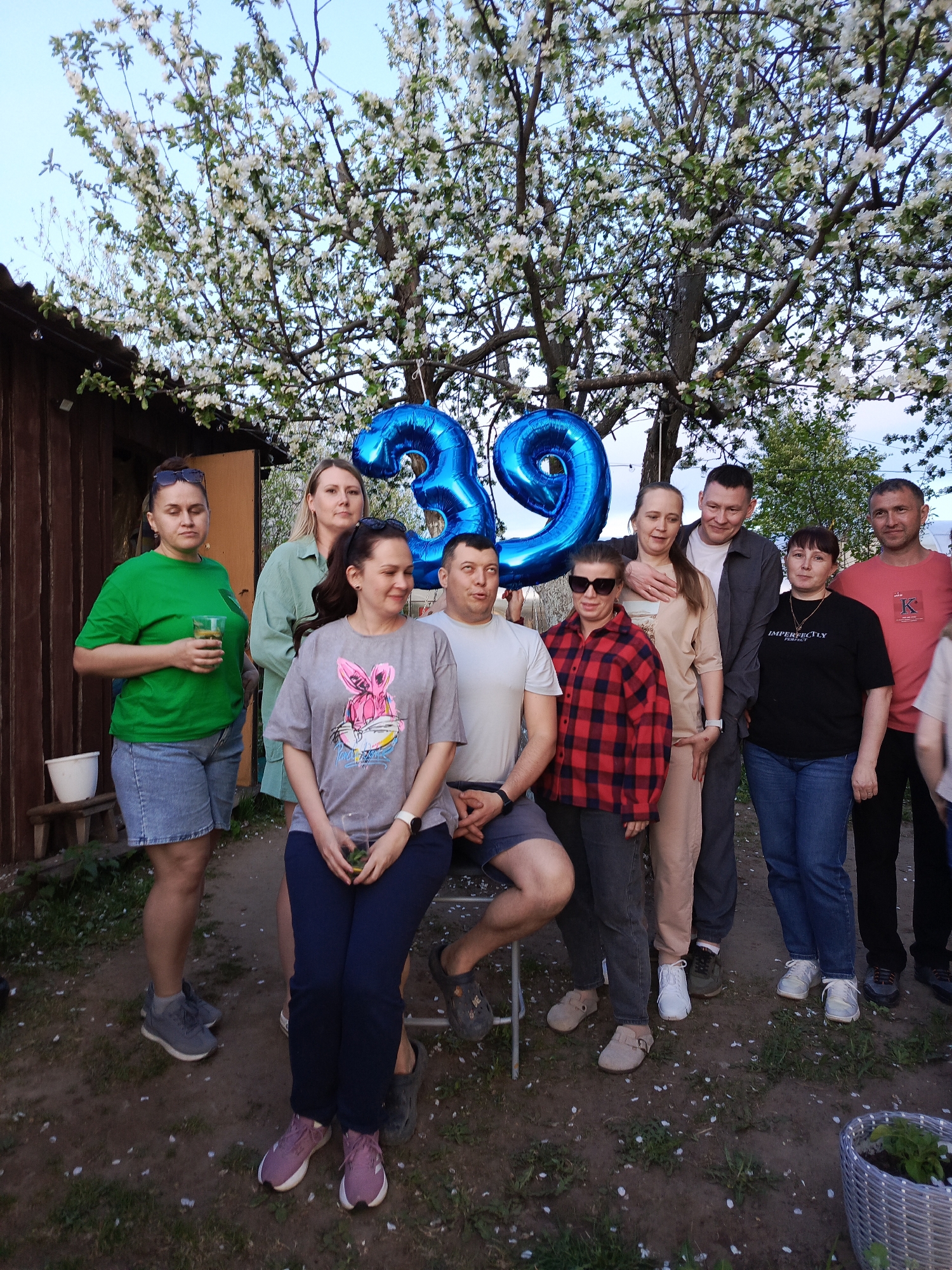 The image shows a group of approximately ten people, possibly family members, gathered outdoors in a backyard setting in an area that is potentially a city in Unknown. The focal point includes large blue "39" balloons, suggesting a 39th birthday celebration. A man in a light-colored t-shirt is sitting on a metal chair. He is being flanked by the balloons.
Several other individuals are standing behind and beside him. They are dressed in a variety of casual clothing. Some of the individuals are holding drinks. A woman in the front is wearing a grey shirt with a pink bunny graphic. A person with sunglasses is also present, wearing a red and black plaid shirt.
The background features a wooden shed or outbuilding on one side, and a tree with white blossoms suggesting springtime in the other, providing depth to the scene. The ground is covered in dirt with scattered patches of grass and white petals.