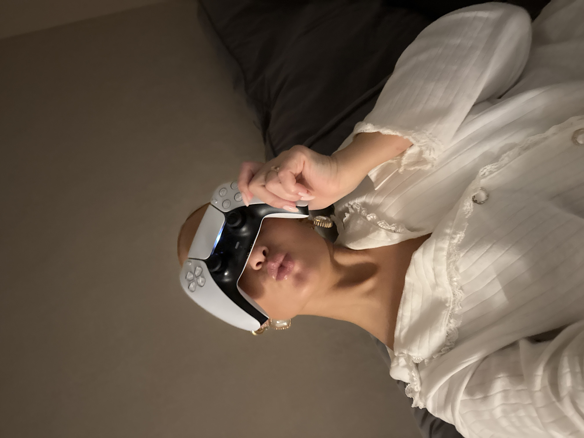 A woman is lying on a bed with a PlayStation 5 controller held up to her face. She is wearing a white blouse with lace trim. The background is a gray pillow and a wall. The location is unspecified in the image.