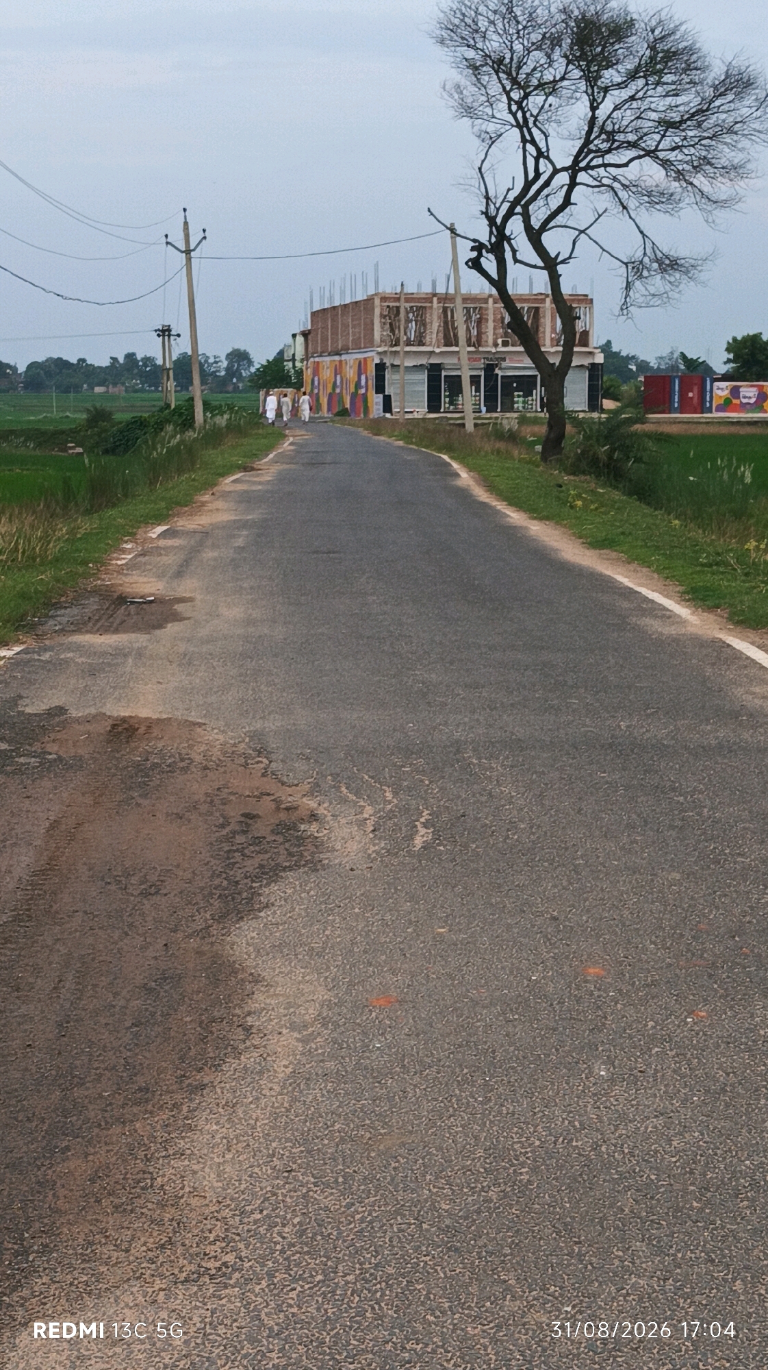A rural road in Barahat, India, stretches into the distance under an overcast sky. On the right side of the road, a building is under construction, with exposed concrete beams and brickwork. The lower facade of the building is adorned with colorful murals. A large, bare tree with intricate branches stands prominently in front of the construction, its silhouette stark against the sky. To the left of the road, lush green fields border the path, with a few electric poles visible in the background. Three individuals, dressed in traditional white Indian attire, are walking away from the camera down the road, towards the building. The road surface itself shows signs of wear and tear, with patches of discolored asphalt and some small debris. A white line marks the edge of the road on the right. The lower right corner of the image displays a timestamp: "31/08/2026 17:04".